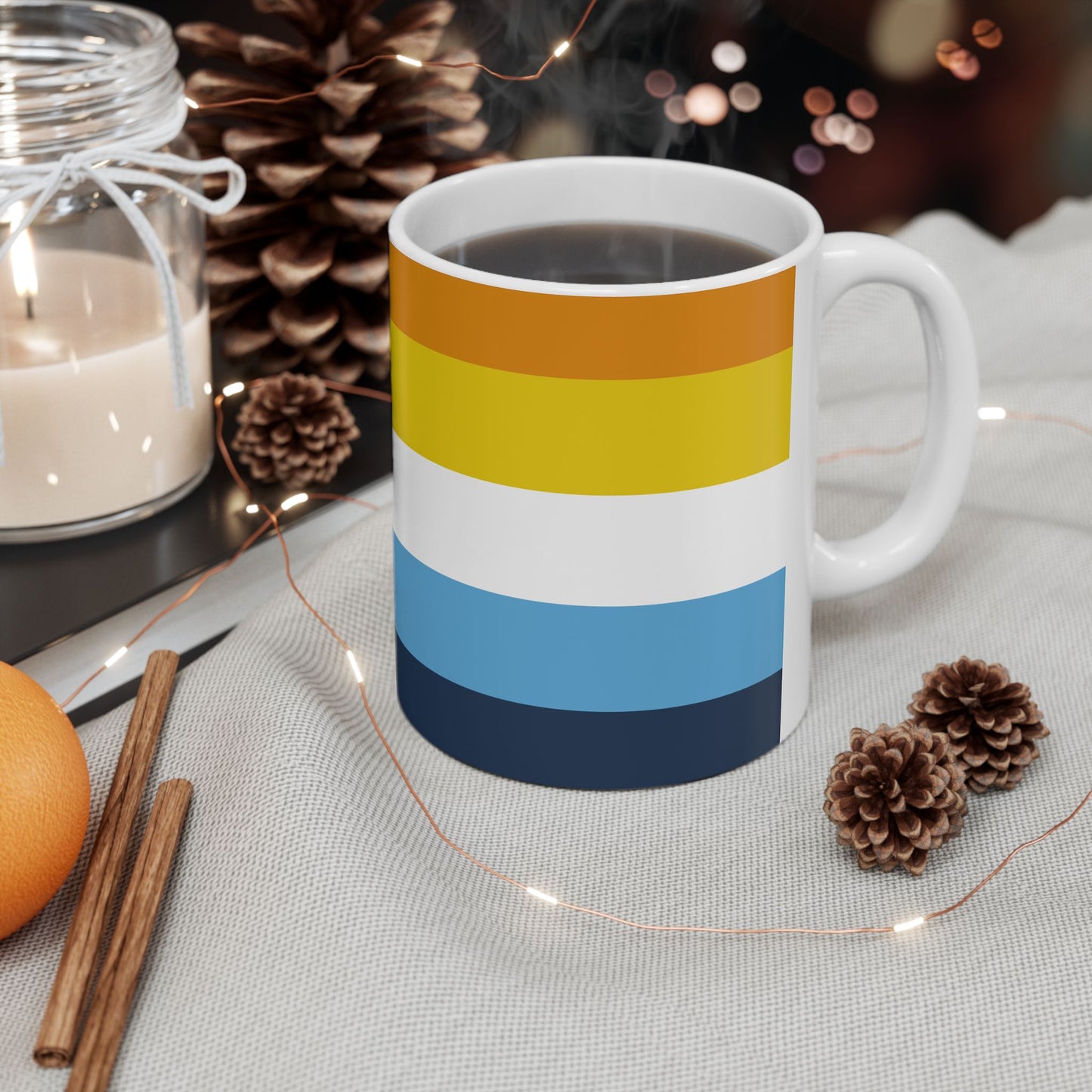 AroAce Pride Flag Mug by WhimSpin - this ceramic mug has colored striped from top to bottom as follows: orange, yellow/gold, white, blue, and navy 11oz size it sits on a clear glass coasger on a white table , near two miniature pinecone, a cinnamon stick and a candle.