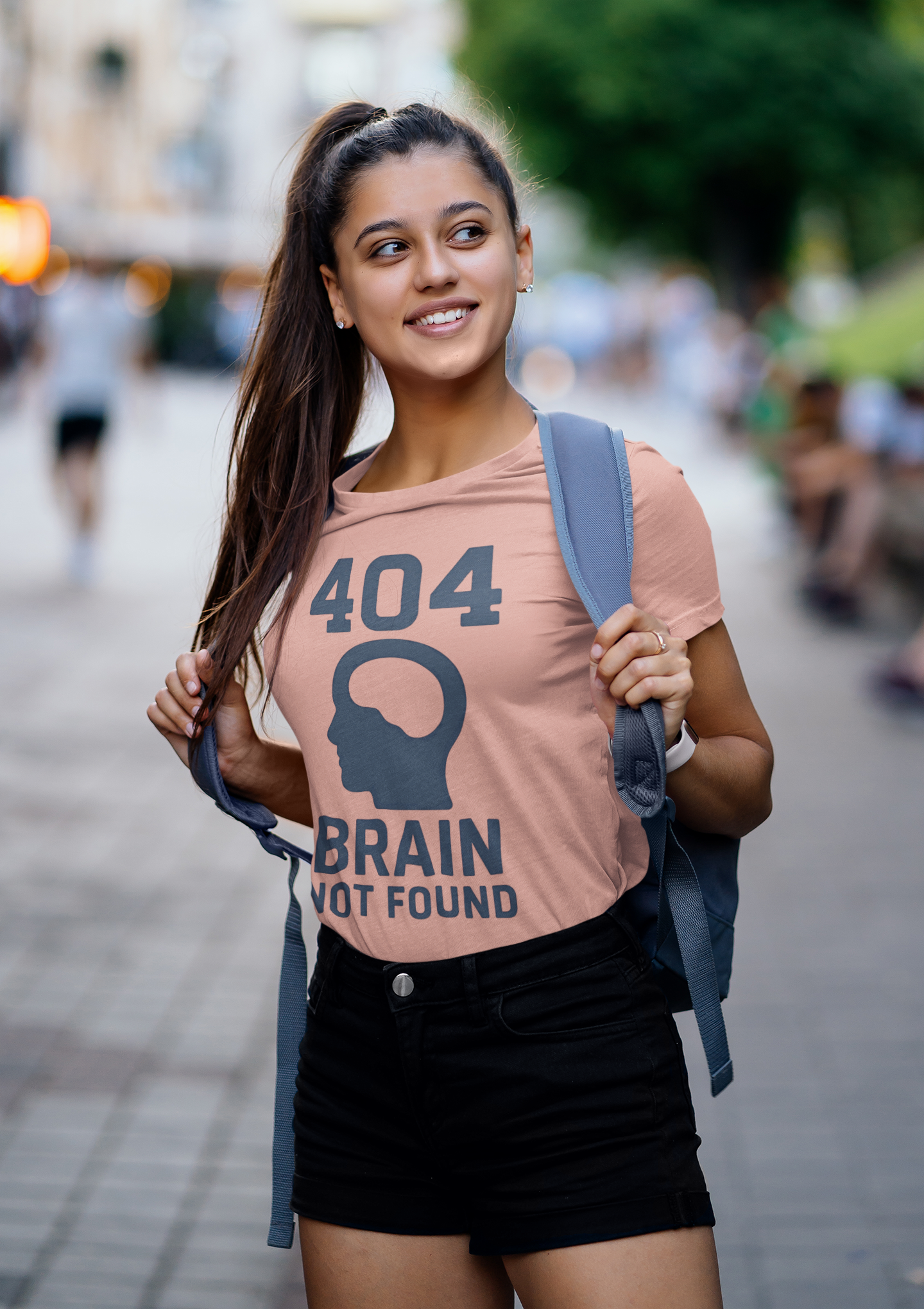 Woman wearing a pink t-shirt a '404 Brain Not Found' from WhimSpin and a backpack on her back on a city street. "404 Brain Not Found T-Shirt" from WhimSpin in gray. The tshirt graphic is a profile image of a human head with an empy hole where the brain should be and the text "404 Brain Not Found."
