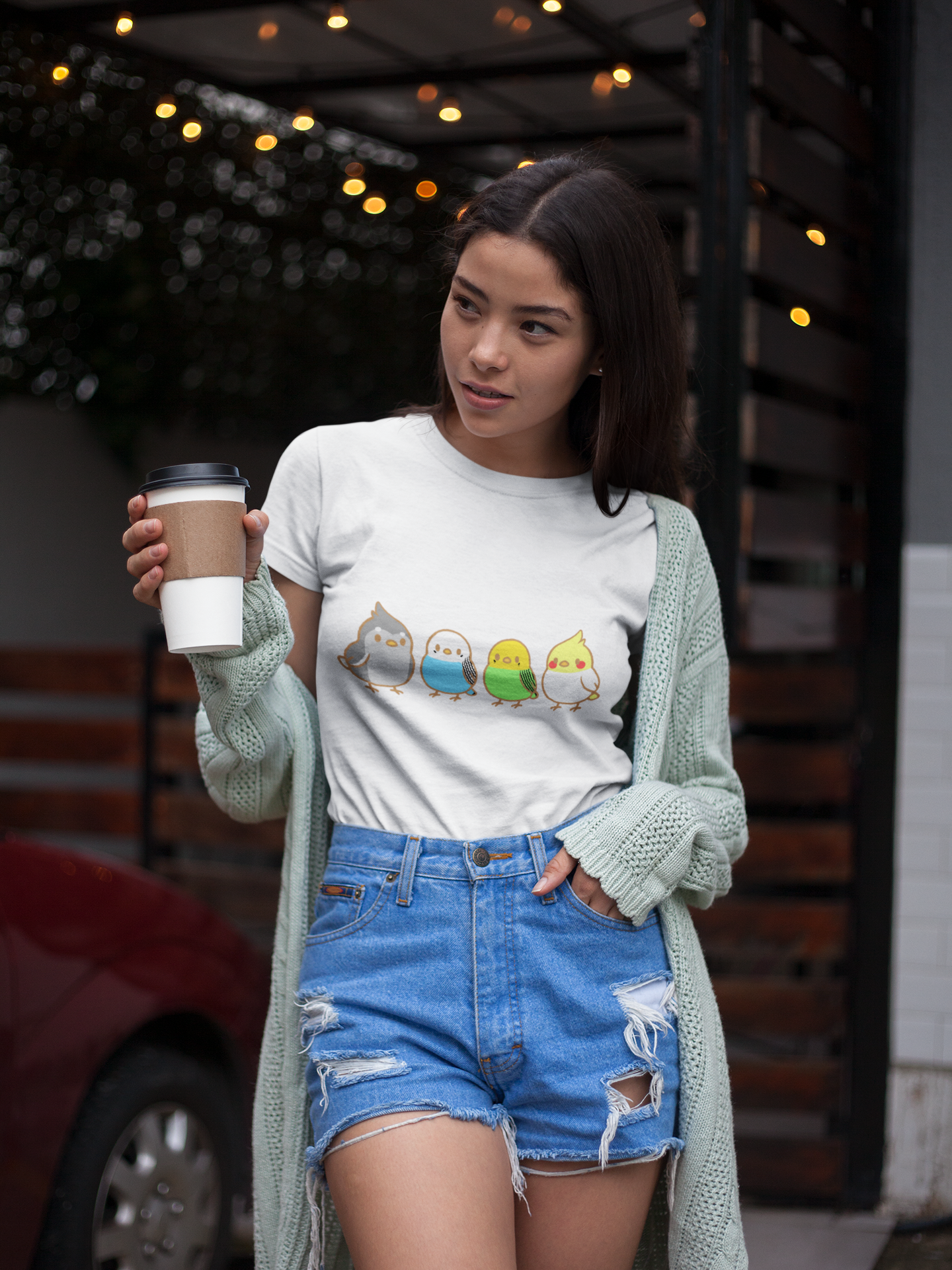 A Young woman holding a cup of coffee wearing a white t-shirt with 4 cartoon pet bird s on it. Chirpy Flock Bird T-Shirt – Cute Tee for Animal Lovers