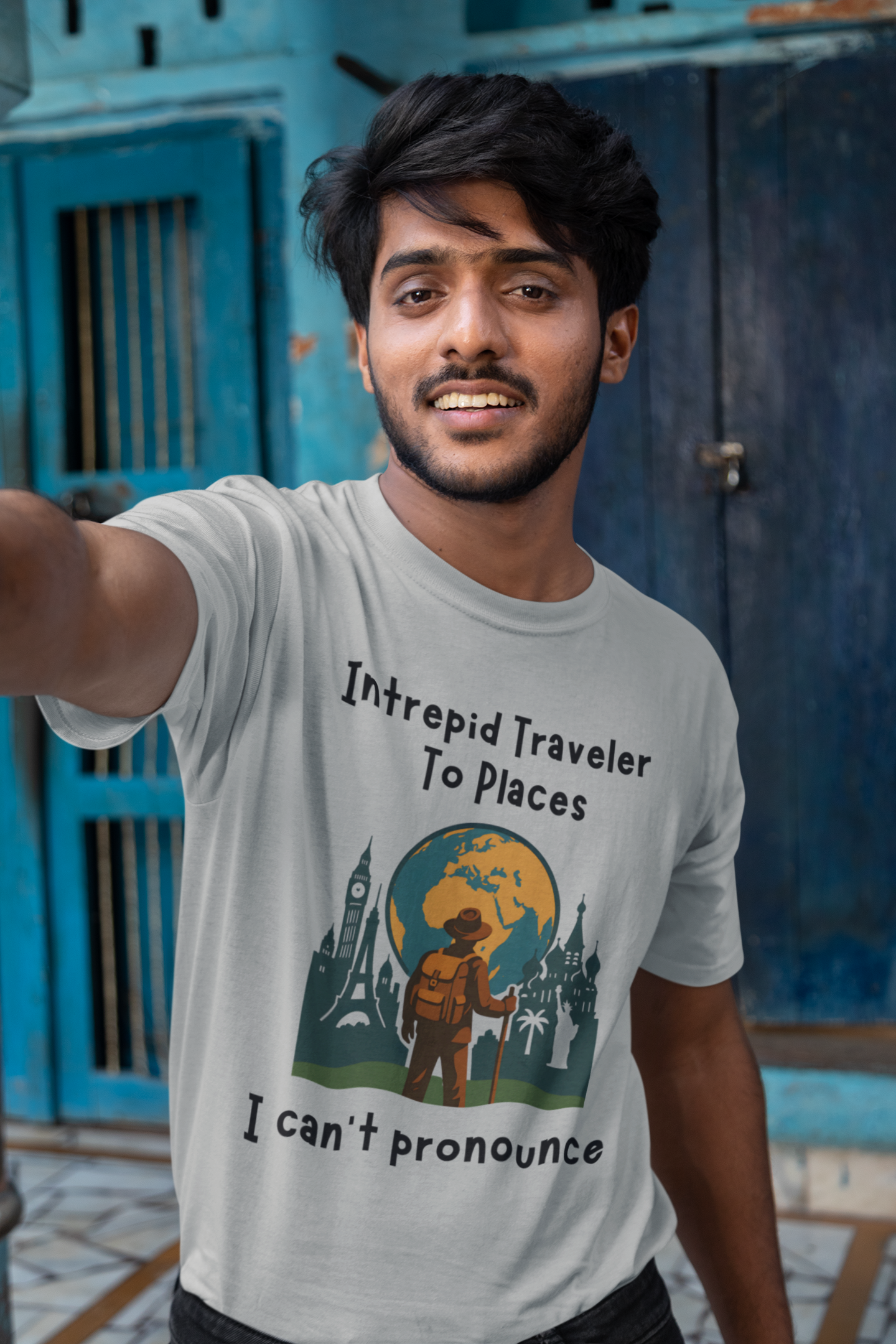 Person wearing a t-shirt with a graphic design of a traveler standing in front of the silohuettes of world landmarks and a globe with the text "Intrepid Traveler to Places I Can't Pronounce" standing in front of a blue door.
