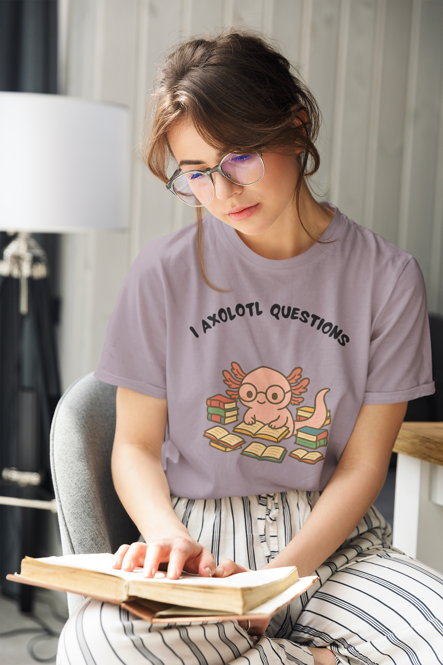 Woman wearing the I Axolotl Questions T-Shirt from WhimSpin in lavender with a graphic design and text, sitting on a chair reading a book.