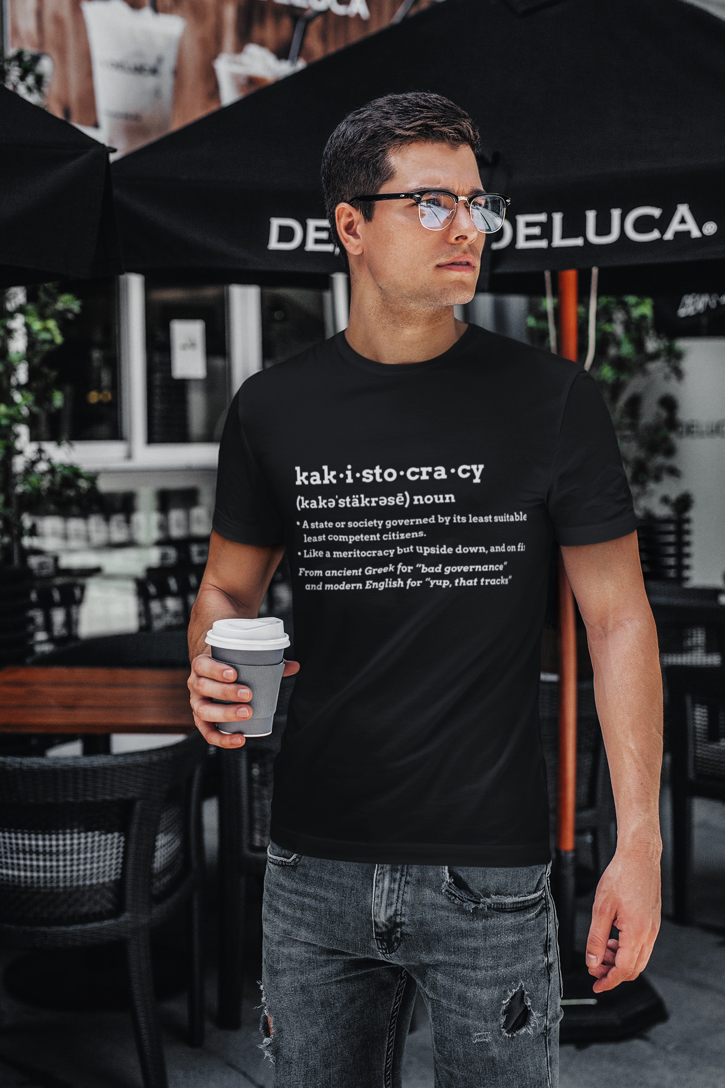 Man wearing the Kakistocracy Definition T-Shirt from WhimSpin in black, holding a coffee cup in an outdoor cafe setting.