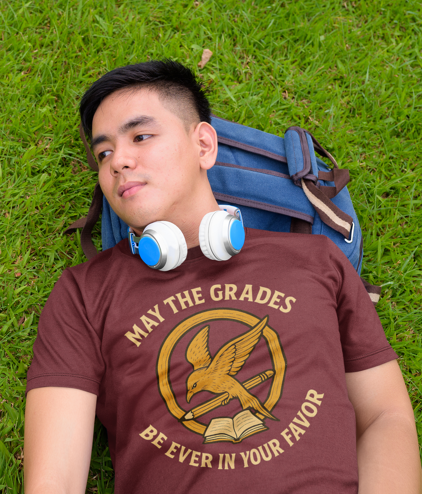 A College age man wears a "May the Grades Be Ever In Your Favor" by WhimSpin in maroon lays on the grass with his head on his backpack like a pillow. The image on the shirt features a parody of the Mockijay design from the voer of the Hunger Games books: a gold circle around a bird in flight carrying a pencil over a open book