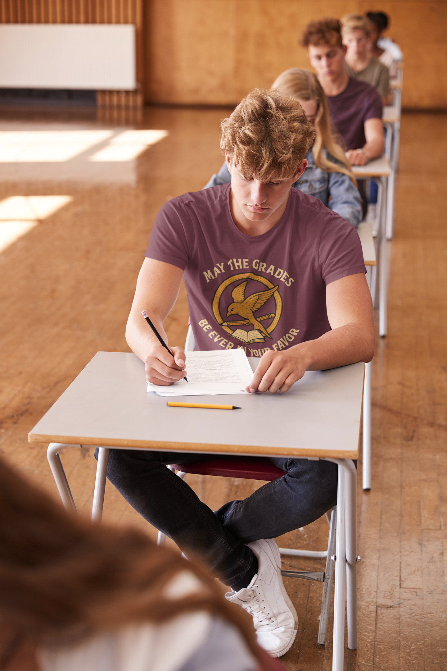 A College age man wears a "May the Grades Be Ever In Your Favor" by WhimSpin in maroon sits at a classroom desk taking a test. The image on the shirt features a parody of the Mockijay design from the voer of the Hunger Games books: a gold circle around a bird in flight carrying a pencil over a open book