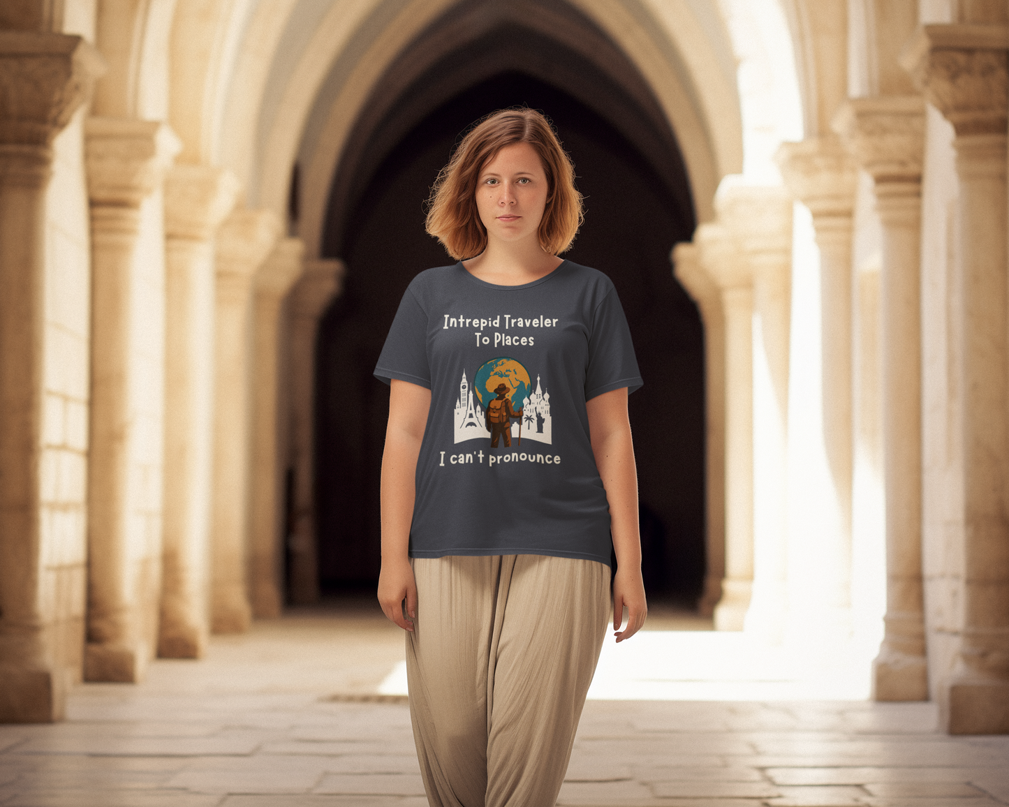 Woman wearing a t-shirt with a graphic design of a traveler standing in front of the silohuettes of world landmarks and a globe with the text "Intrepid Traveler to Places I Can't Pronounce" in an architectural setting