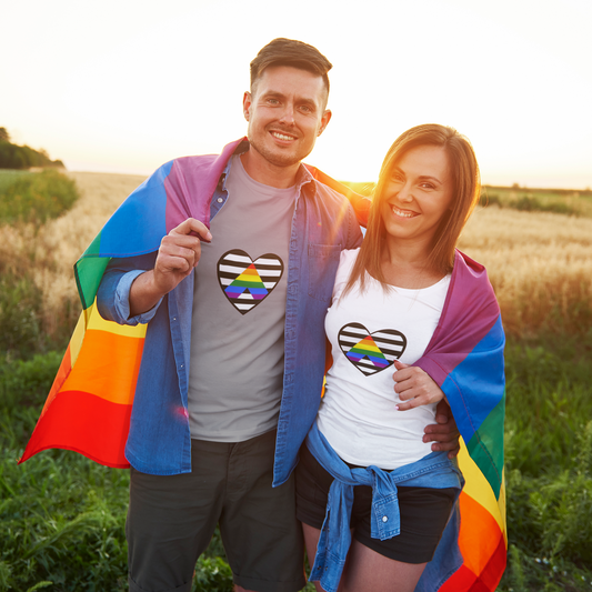 Heart Ally Flag T-Shirt – Subtle LGBTQ+ Support with Bold Pride worn by a smiling couple wrapped in rainbow flags at sunset, featuring the straight ally heart symbol on matching shirts during an outdoor pride event.