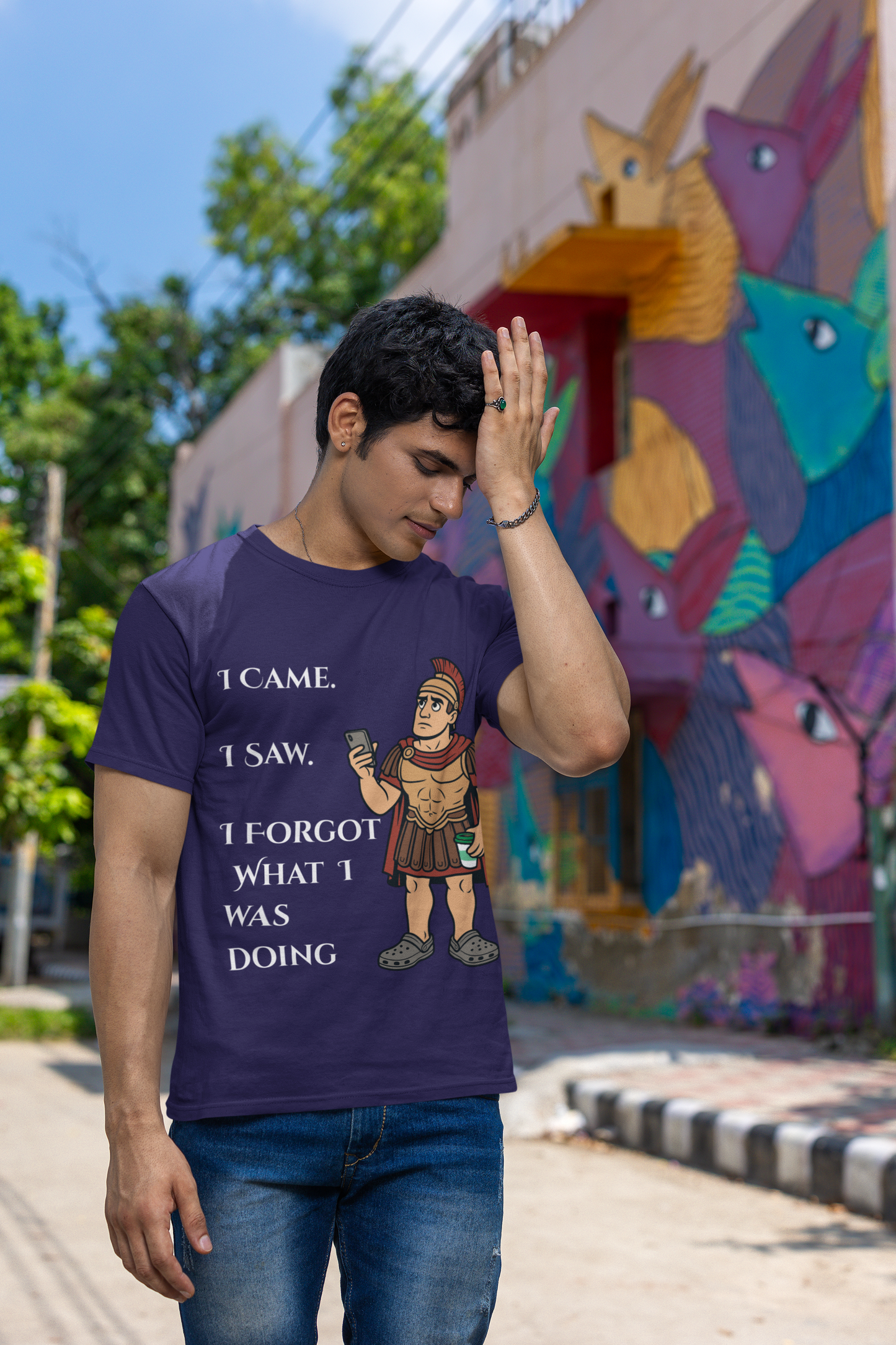 a man wearing a team navy I Came, I Saw, I Forgot T-Shirt – Funny ADHD t shirt from WhimSpin with text and graphics, standing in front of a colorful mural. he has his hand raised to his forehead as if he is just remembering something he forgot.