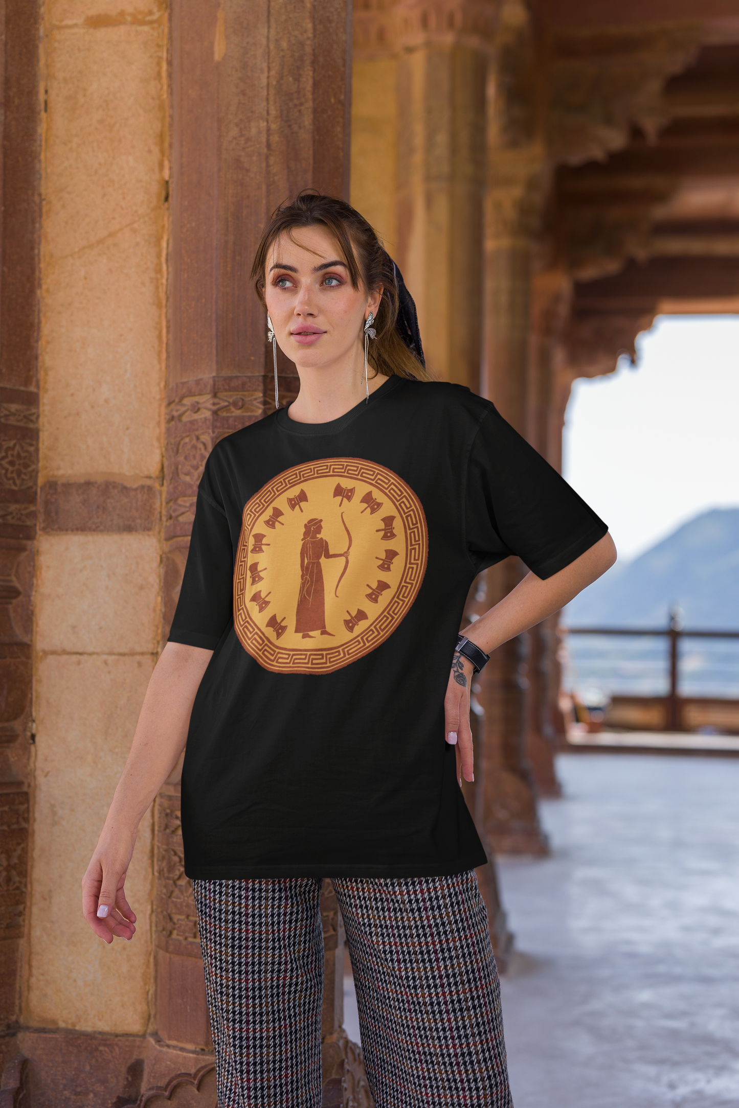 A woman with dark hair stands with her left hand on her hip. She stands in an ancient building and is wearing the Penelope Epic Queen of Ithaca T shirt in black from Whimspin. The shirt features an image of a woman in ancient Grecian robes holding a bow and surrounded by 12 axes. The images is in terracotta cotta red and gold in the style of ancient amphora art.