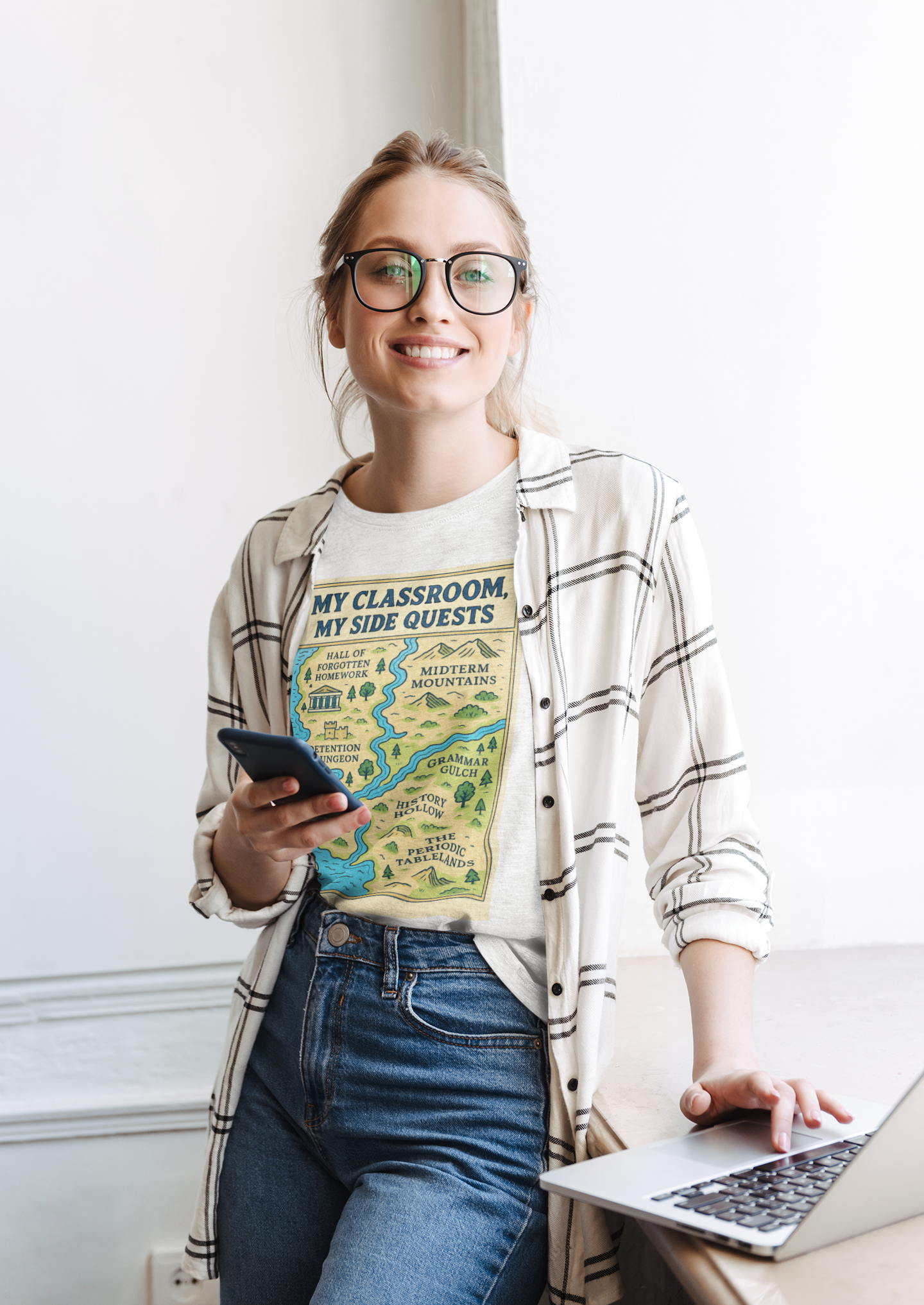 A Woman wearing a the "My Classroom, My Side Quests T-Shirt" from WhimSpin leans on a table with her hand on a laptop. The t-shirtdesign features a maptitled "MyClassroom, My Sidequest" and has classroom themed landmarks.