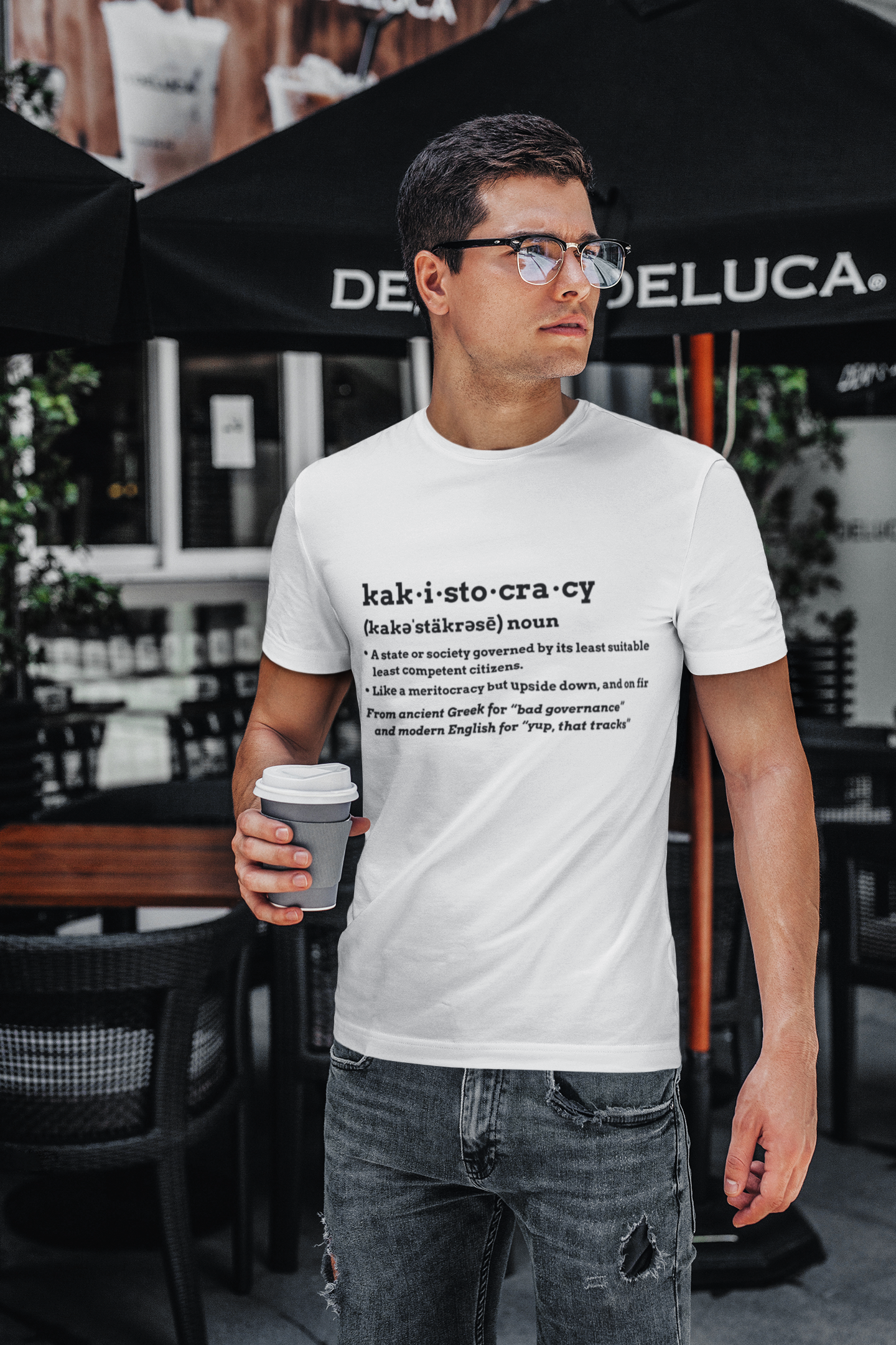 Man wearing a white kakistocracy definition t-shirt from WhimSpin, holding a coffee cup in an outdoor cafe setting.