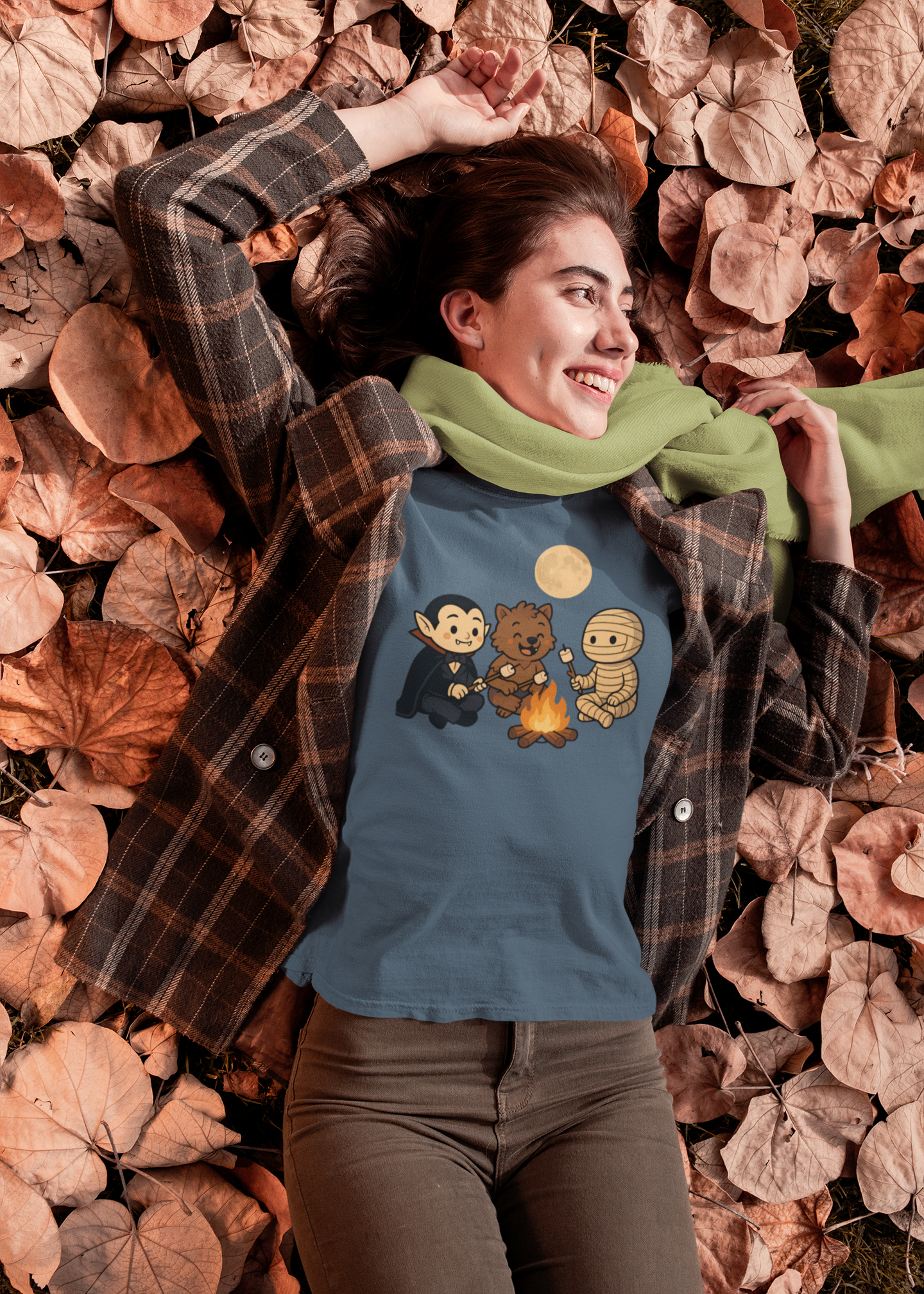 Woman lying on autumn leaves in a green scarf and plaid jacket wearing the Heather Deep Teal Halloween Monsters Campfire T-Shirt by WhimSpin, featuring a cute vampire, werewolf, and mummy roasting marshmallows under a full moon.