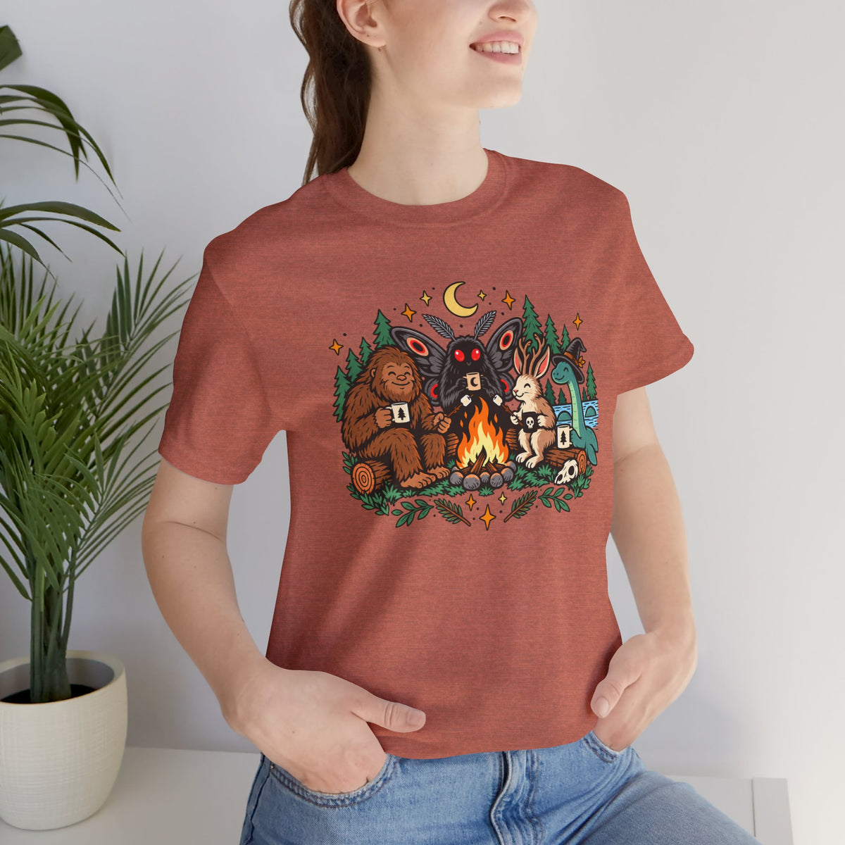 Rust crew-neck campfire cryptid tee with colorful front graphic, worn with jeans