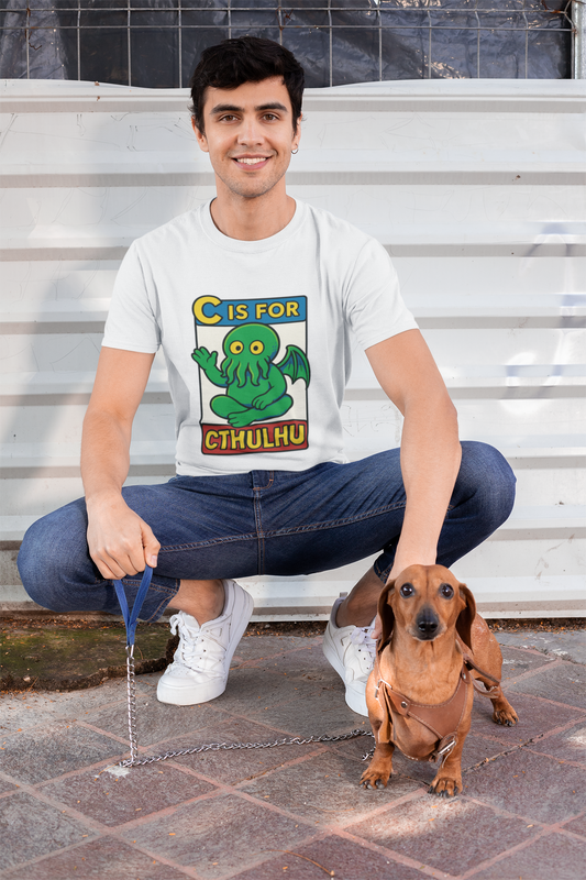 Man wearing white funny Cthulhu shirt, "C is for Cthulhu" tshirt, and blue jeans squats with his dachshund infant of a white wall