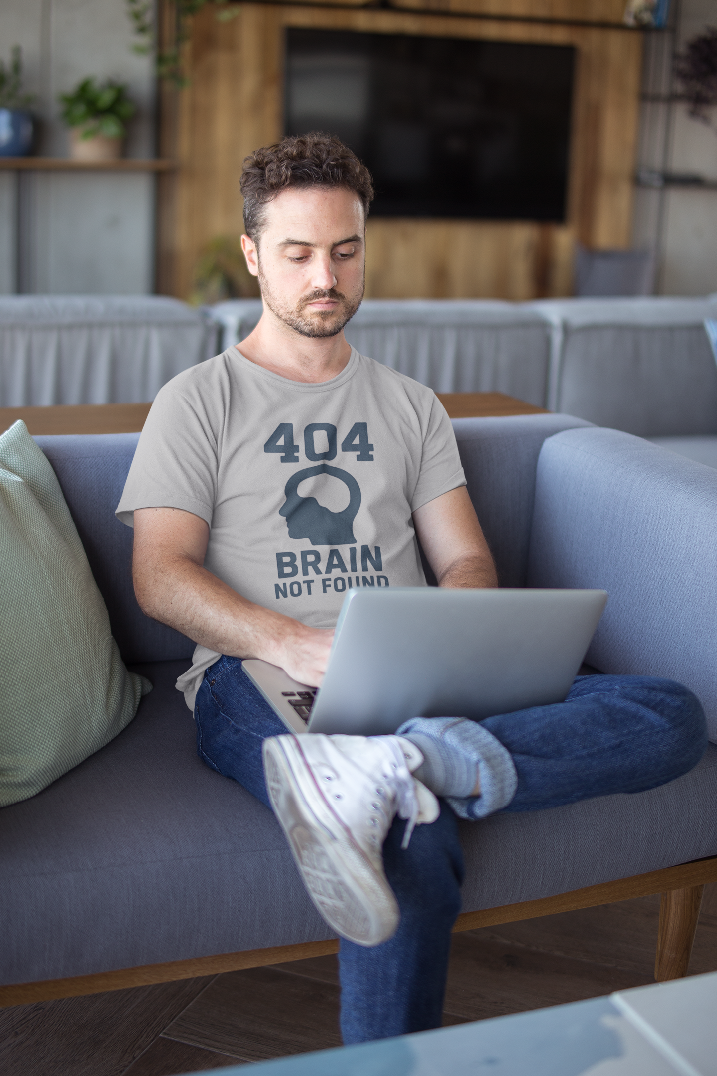 Man sitting on a couch using a laptop in a living room wears the "404 Brain Not Found T-Shirt" from WhimSpin in gray. The tshirt graphic is a profile image of a human head with an empy hole where the brain should be and the text "404 Brain Not Found."