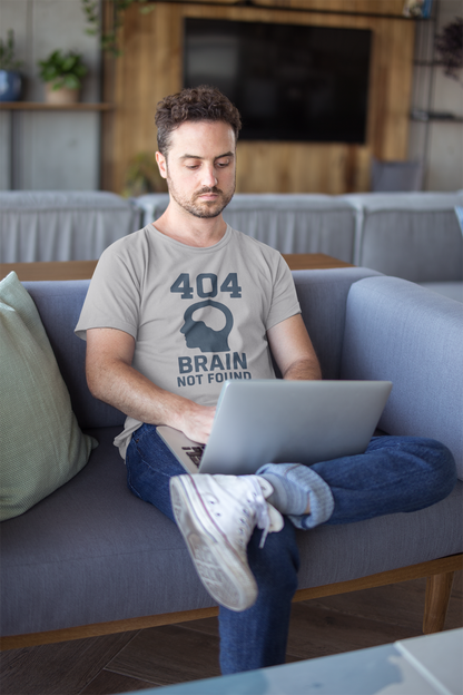 Man sitting on a couch using a laptop in a living room wears the "404 Brain Not Found T-Shirt" from WhimSpin in gray. The tshirt graphic is a profile image of a human head with an empy hole where the brain should be and the text "404 Brain Not Found."