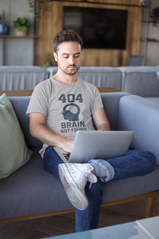 Man sitting on a couch using a laptop in a living room wears the "404 Brain Not Found T-Shirt" from WhimSpin in gray. The tshirt graphic is a profile image of a human head with an empy hole where the brain should be and the text "404 Brain Not Found."