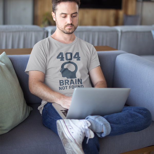 404 Brain Not Found T-Shirt – Silver tee featuring a bold 404 error code graphic, worn by a man sitting cross-legged on a grey couch with a laptop, perfect tshirt for techies working remote or experiencing a brain fart moment