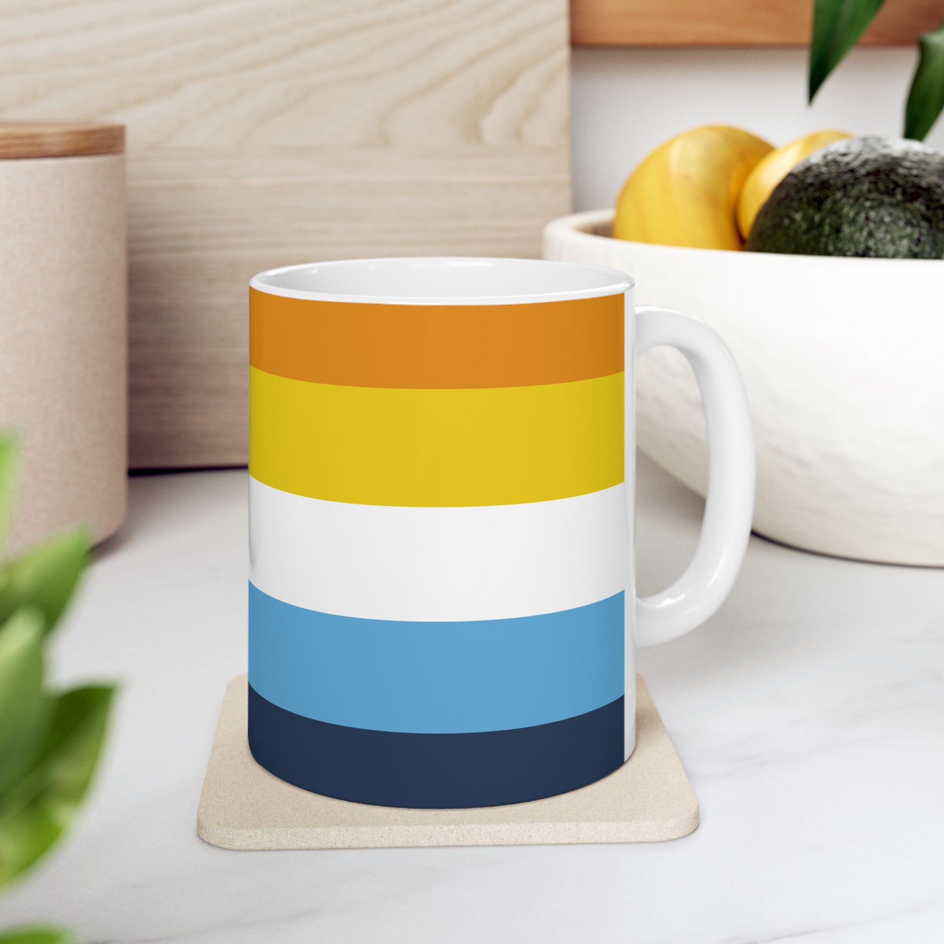 AroAce Pride Flag Mug by WhimSpin sits on a coaster on a white surface near a bowl of fruit and a plant. The ceramic mug has colored striped from top to bottom as follows: orange, yellow/gold, white, blue, and navy 