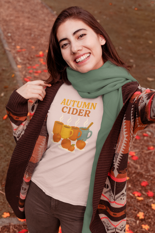 Autumn Cider T-Shirt – Cozy Fall Graphic Tee with Steaming Mugs in Heather Peach Prismvworn by a smiling woman in a fall outfit and sage green scarf, surrounded by autumn leaves