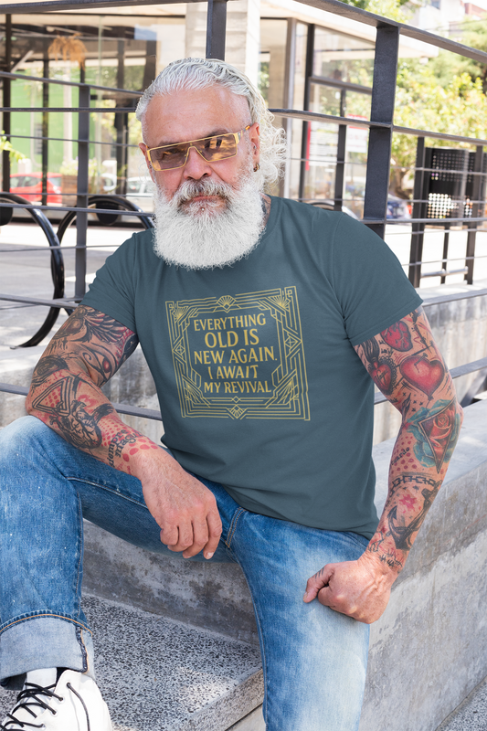 Bearded man with tattoos and sunglasses sits on concrete steps wearing the Everything Old Is New Again – I Await My Revival shirt in deep teal, featuring a vintage gold Art Deco frame.