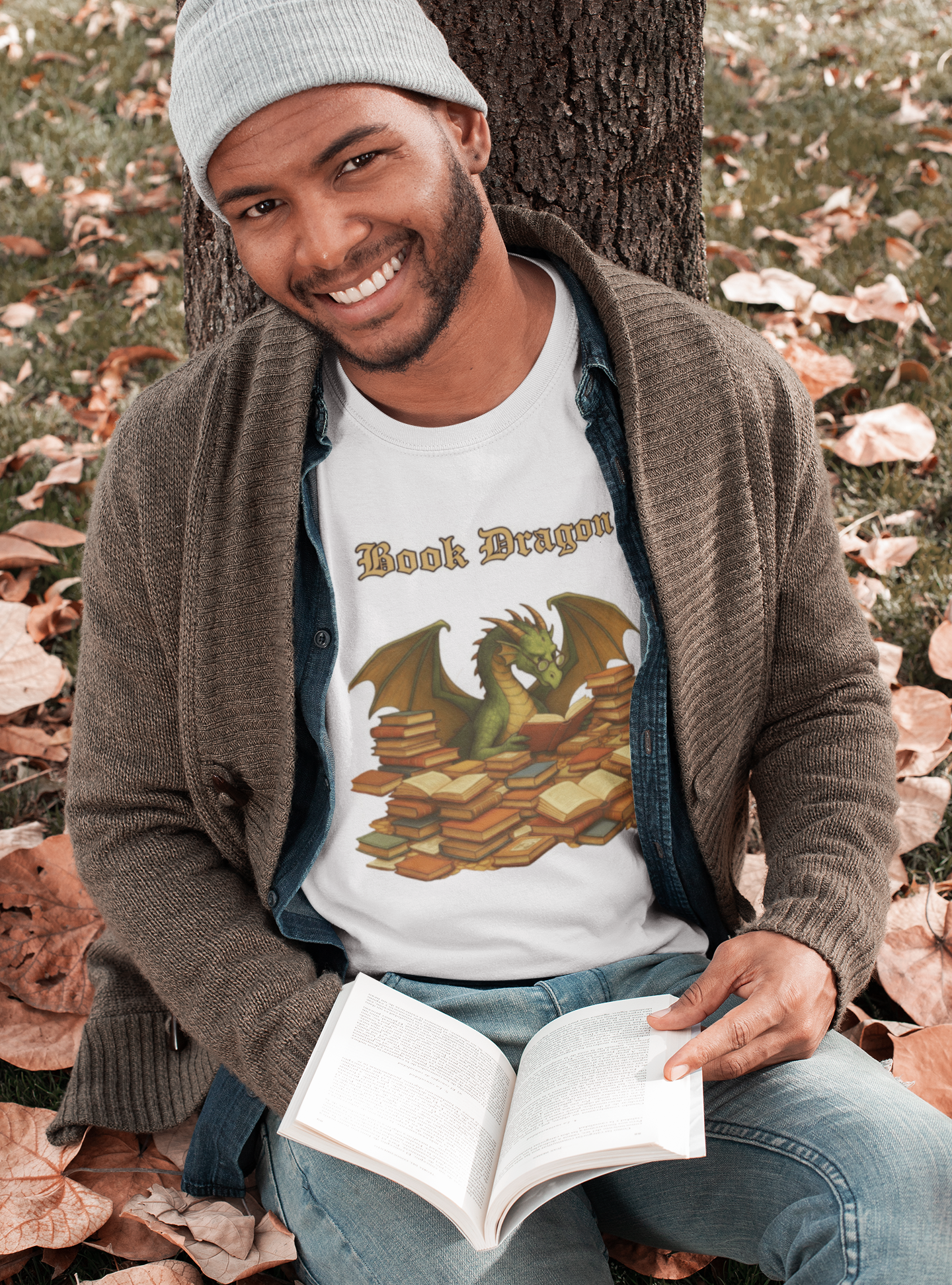 Book Dragon T-Shirt – Funny Book Lover Shirt featuring a smiling man in a gray beanie and cardigan reading under a tree in autumn