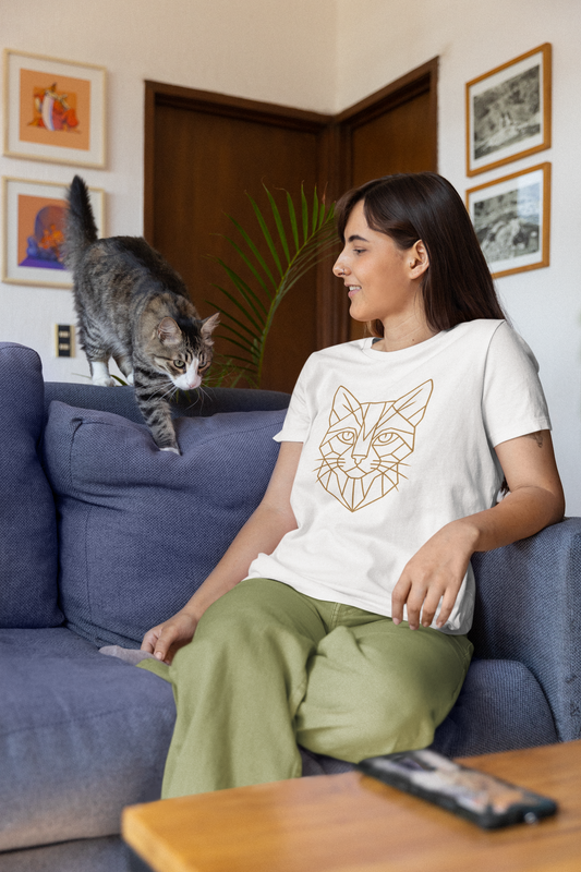A dark haired woman wearing the Geometric Cat T-Shirt – Minimalist Line Art Teefrom WhimSpin in white and a lime green pants sits on a blue sofa and looks at an approaching tabby cat.