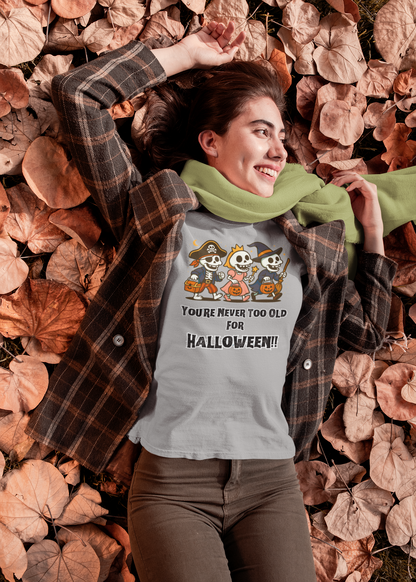 Smiling woman in a light grey (silver) version of the Never Too Old for Halloween T-Shirt – Funny Adult Halloween Shirt; skeleton trio design pops in the autumn-themed setting with cozy plaid layers and scarf.