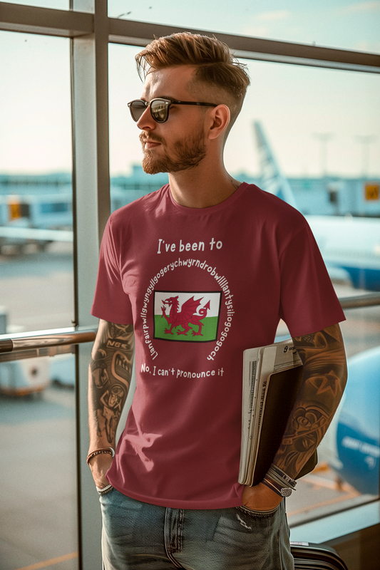 A man wearing the Llanfairpwllgwyngyllgogerychwyrndrobwllllantysiliogogogoch T-Shirt – Funny Wales Travel Merch in canvas red at an airport gate, with tattoos and sunglasses, holding a newspaper beside a large window.