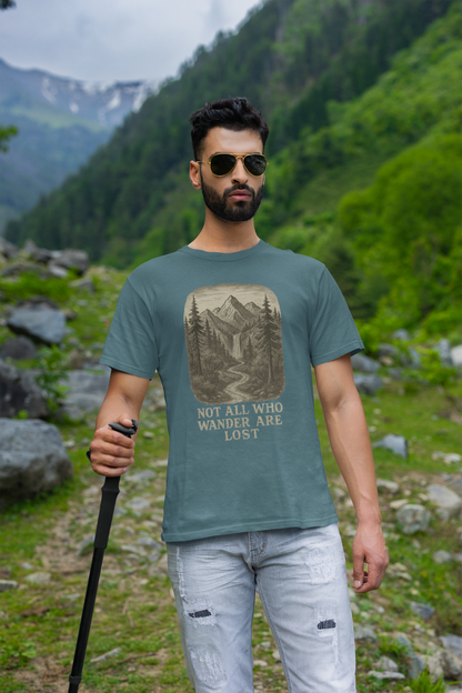 Man in sunglasses hiking a rocky mountain trail in heather deep teal “Not All Who Wander Are Lost T-Shirt – Tolkien Travel Tee for Nature Lovers” with scenic waterfall and forest graphic