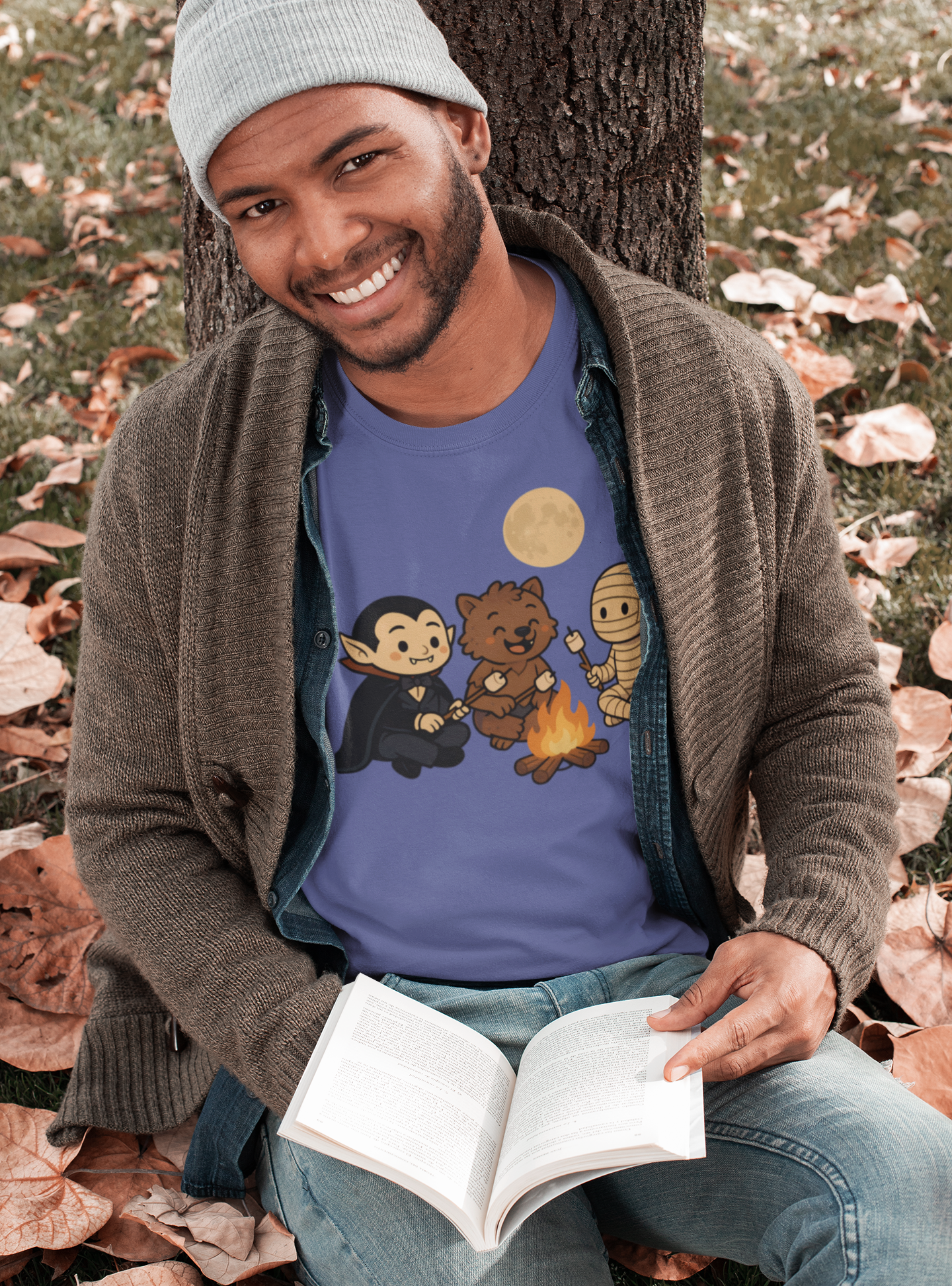 Man sitting on autumn leaves by a tree, smiling while reading a book and wearing the Heather Team Purple Halloween Monsters Campfire T-Shirt by WhimSpin, which shows a vampire, werewolf, and mummy roasting marshmallows.
