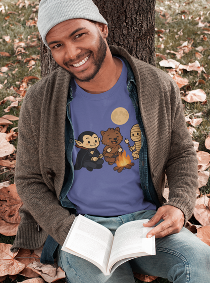 Man sitting on autumn leaves by a tree, smiling while reading a book and wearing the Heather Team Purple Halloween Monsters Campfire T-Shirt by WhimSpin, which shows a vampire, werewolf, and mummy roasting marshmallows.