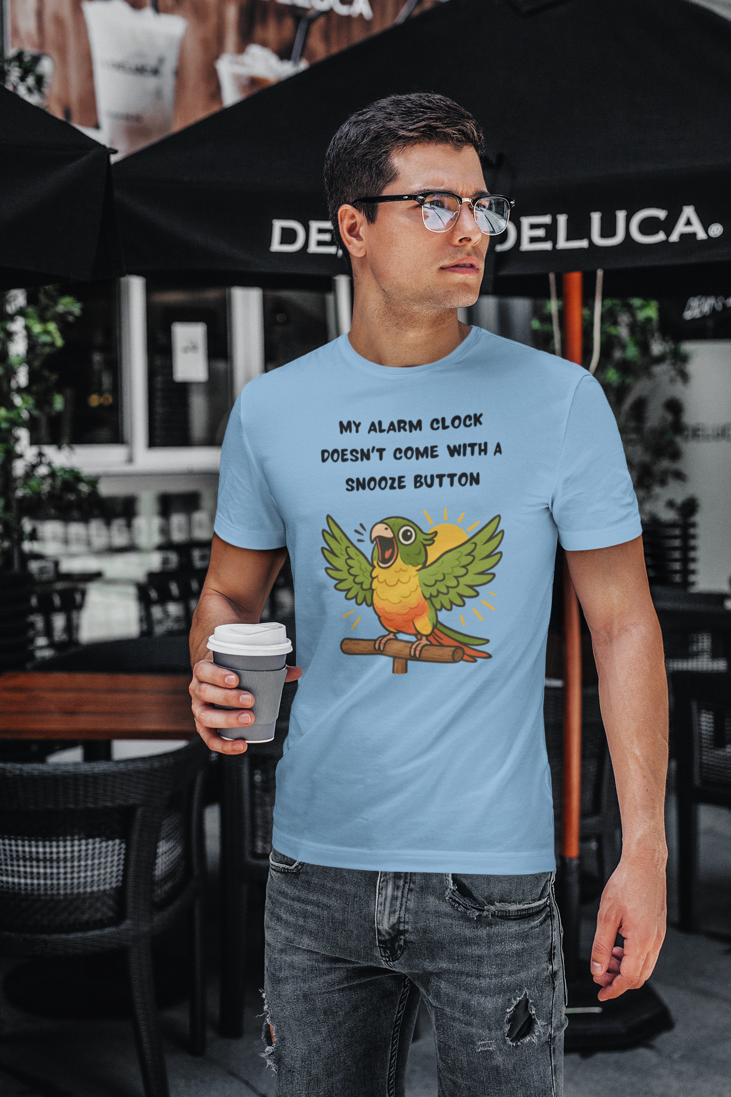 A man wearing the Parrot Alarm Clock T-Shirt – Funny Parrot Wake-Up Call Tee in Ocean Blue in a city café setting, holding a coffee cup and displaying the bold, colorful parrot illustration and playful wake-up call design.