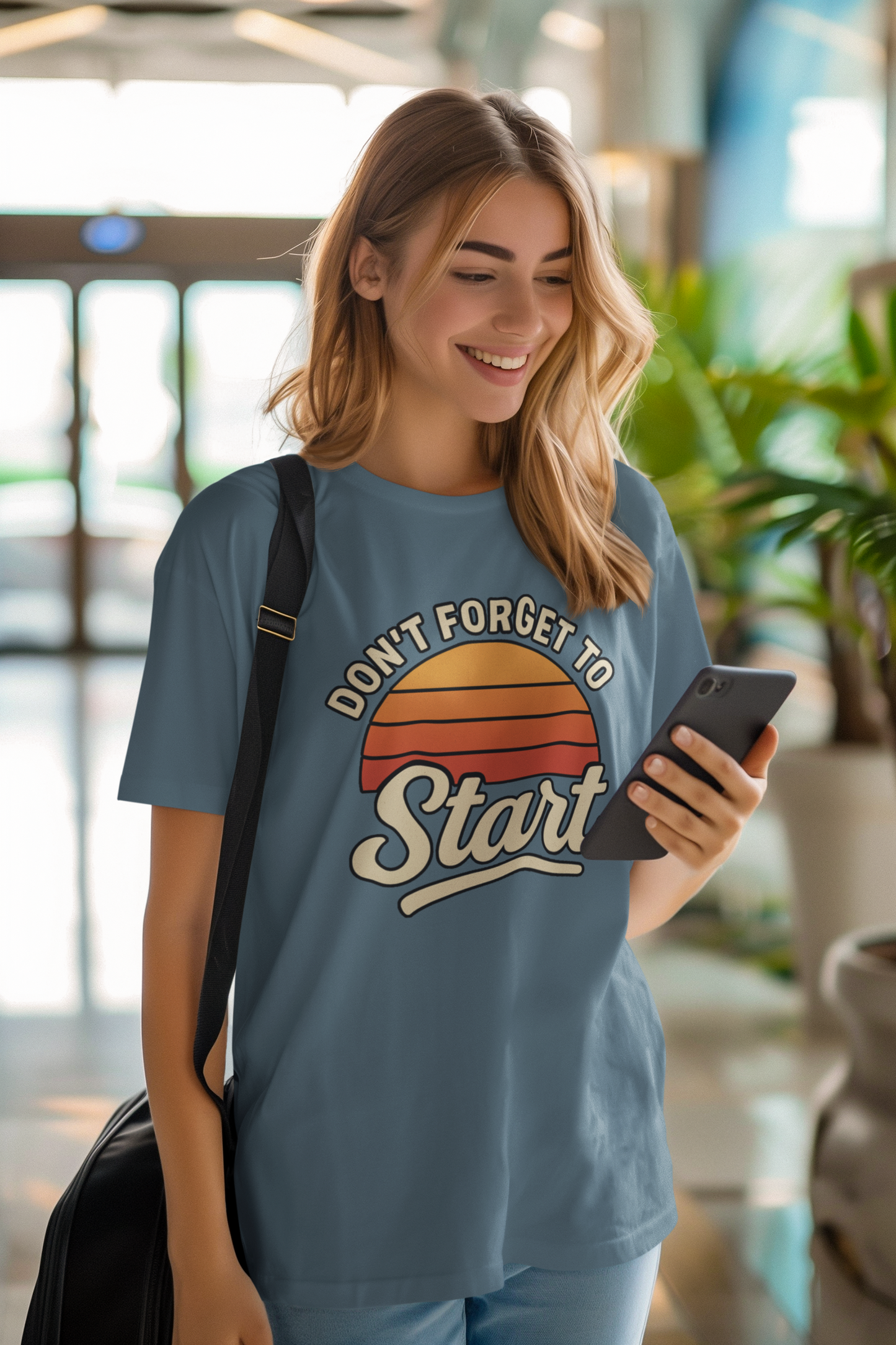 Don’t Forget to Start T-Shirt – Motivational Graphic Tee for Dreamers shown on woman walking indoors smiling, with crossbody bag and smartphone