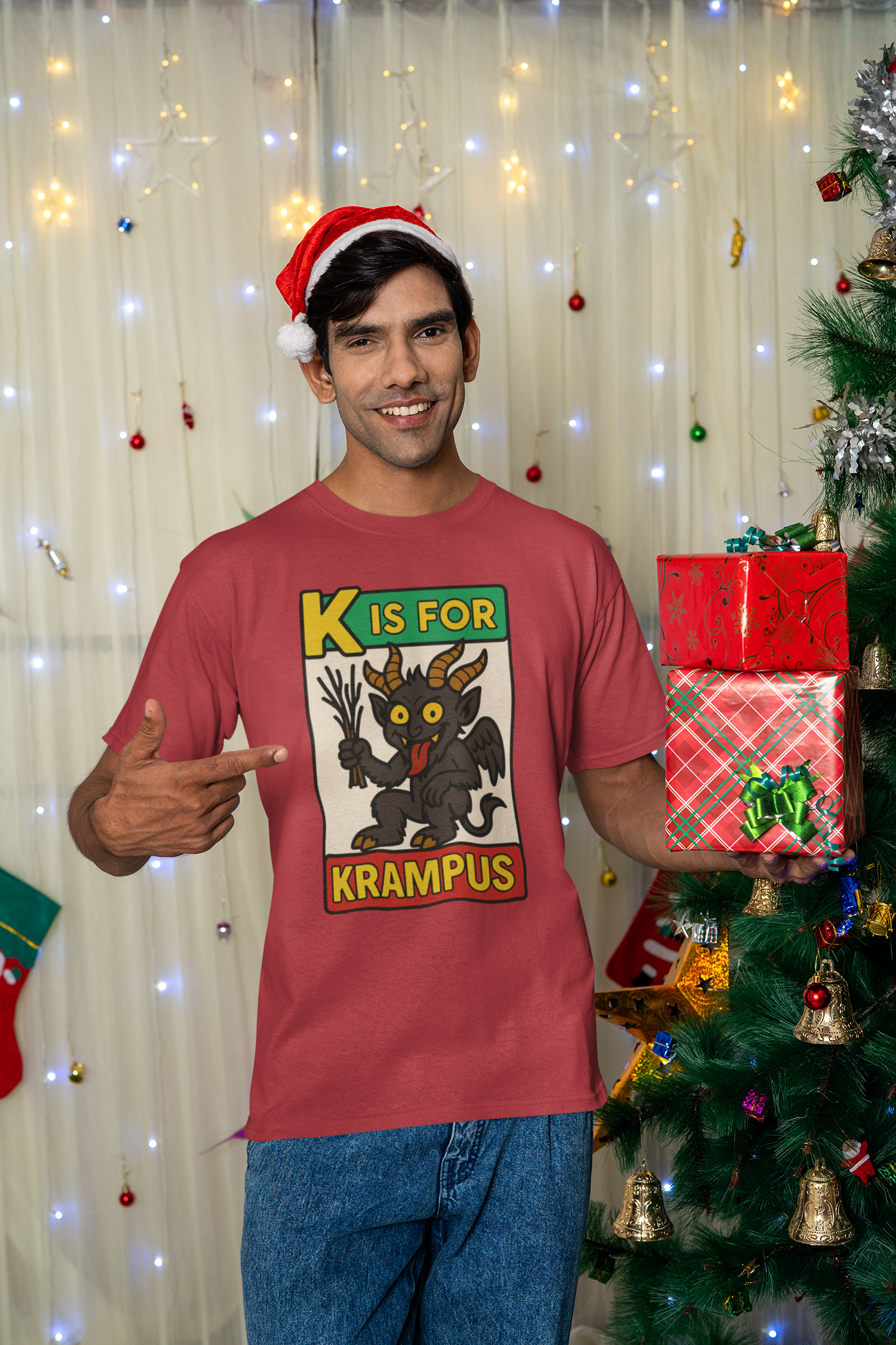 K is for Krampus Alphabet Holiday Graphic T-Shirt in Canvas Red worn by man smiling beside a decorated Christmas tree