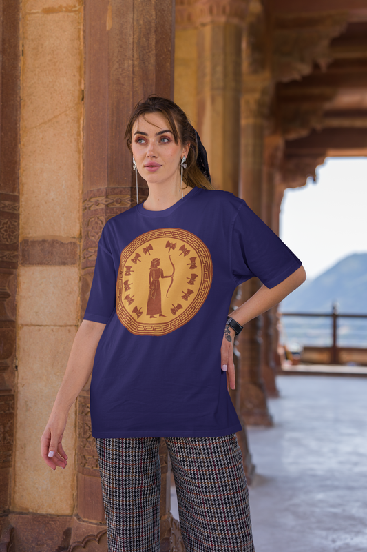 A woman with dark hair stands with her left hand on her hip. She stands in an ancient building and is wearing the Penelope Epic Queen of Ithaca T shirt from Whimspin. The shirt features an image of a woman in ancient Grecian robes holding a bow and surrounded by 12 axes. The images is in terracotta cotta red and gold in the style of ancient amphora art. 
