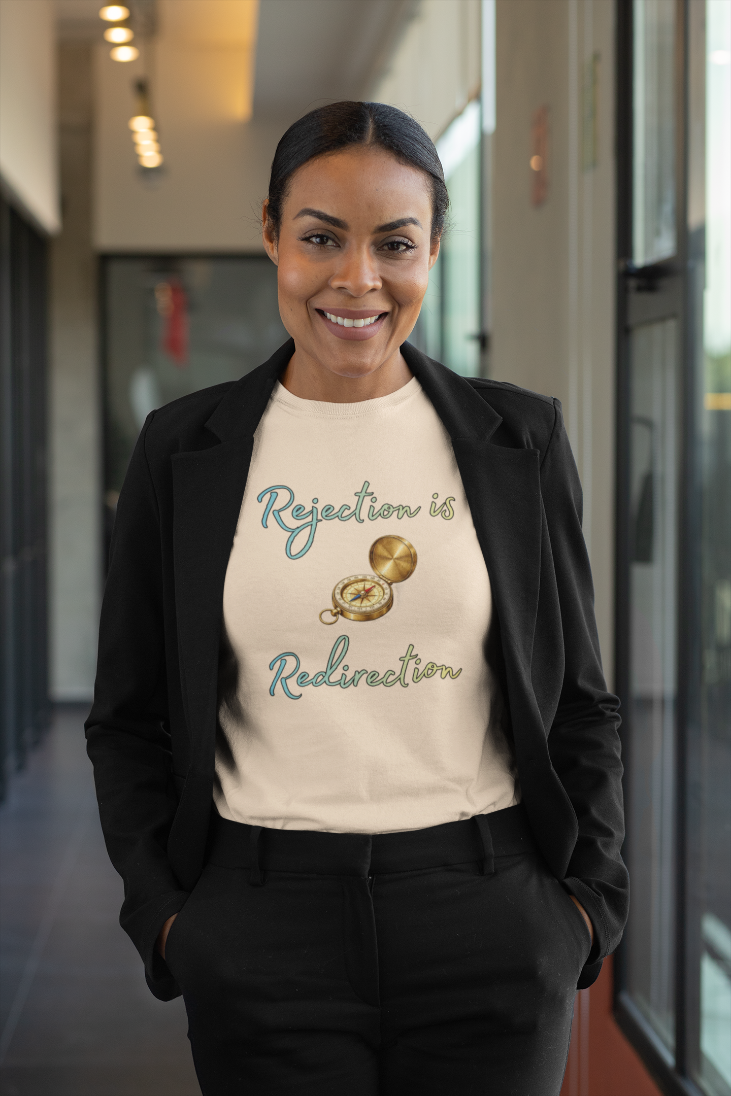 Rejection Is Redirection T-Shirt – Motivational Compass Quote Tee in heather sand dune worn with a tailored black suit in an indoor corridor, highlighting the compass graphic and redirection message