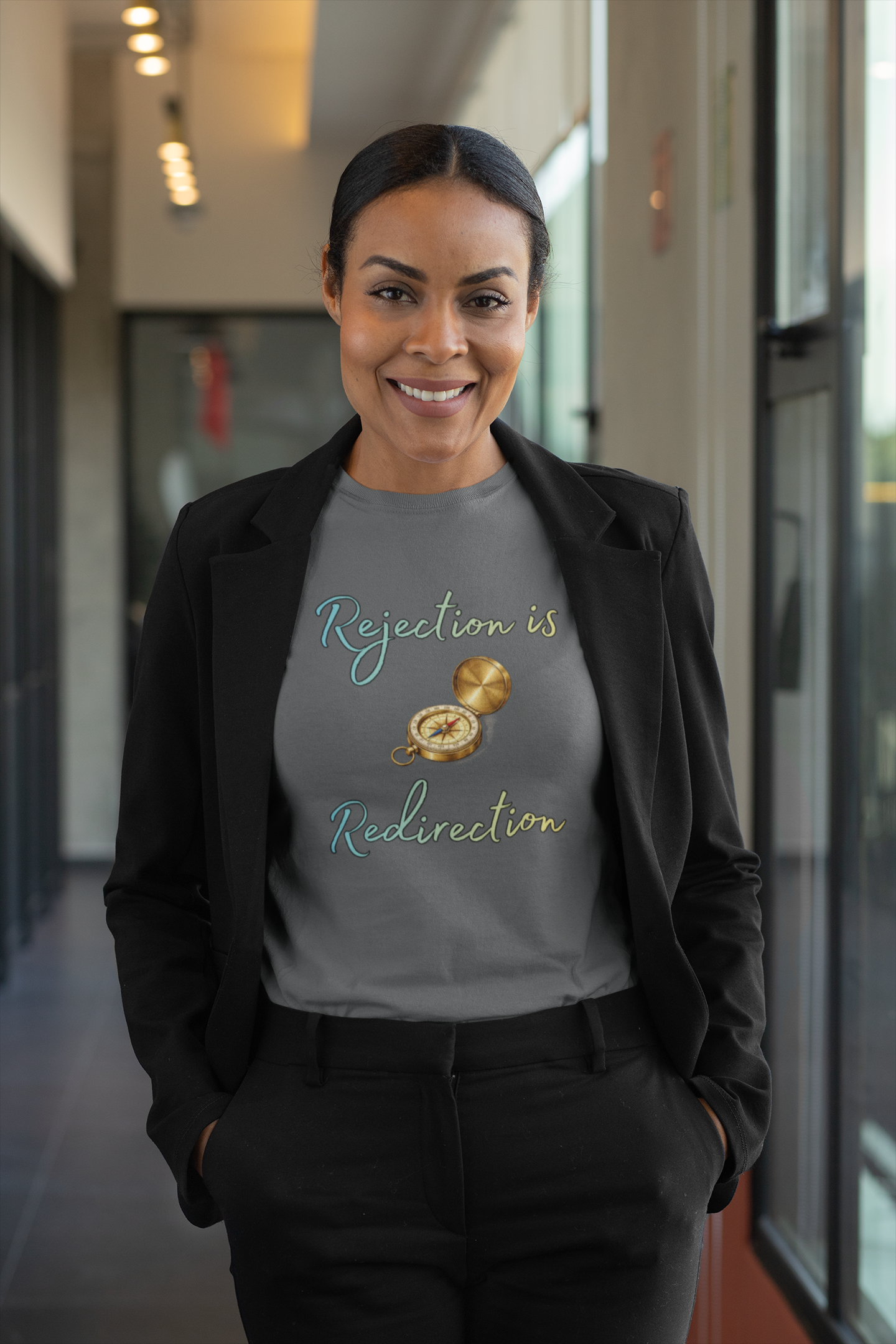 Rejection Is Redirection T-Shirt – Motivational Compass Quote Tee in asphalt gray worn with a black blazer in a bright office hallway, showing the rejection is redirection quote and compass illustration
