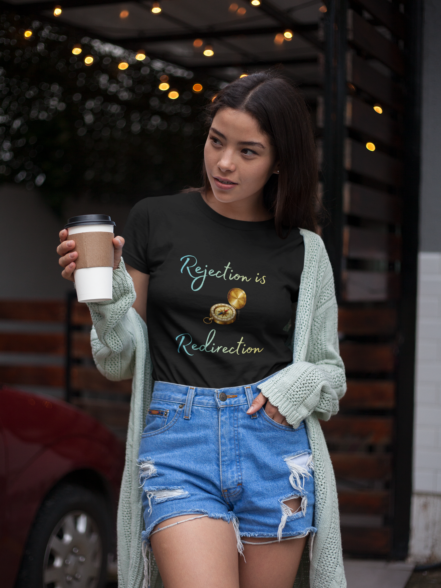 Rejection Is Redirection T-Shirt – Motivational Compass Quote Tee worn by a woman holding a takeaway coffee outdoors under string lights, styled with a mint cardigan and denim shorts, featuring the rejection is redirection compass graphic