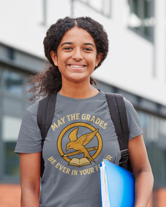 A college aged woman wears a "May the Grades Be Ever In Your Favor" by WhimSpin in slate blue stands with a backpack on her back and a blue binder under her left arm.  The image on the shirt features a parody of the Mockijay  design from the voer of the Hunger Games books: a gold circle around a bird in flight carrying a pencil over a open book. 