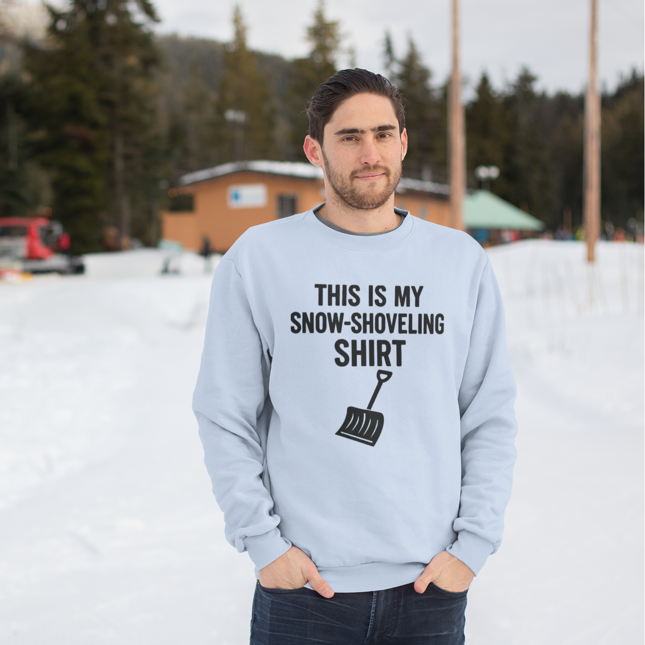 This Is My Snow-Shoveling Shirt – Funny Winter Chore Sweatshirt worn by a man standing outdoors in a snowy winter setting with cabins in the background, featuring the snow shovel graphic and bold winter humor