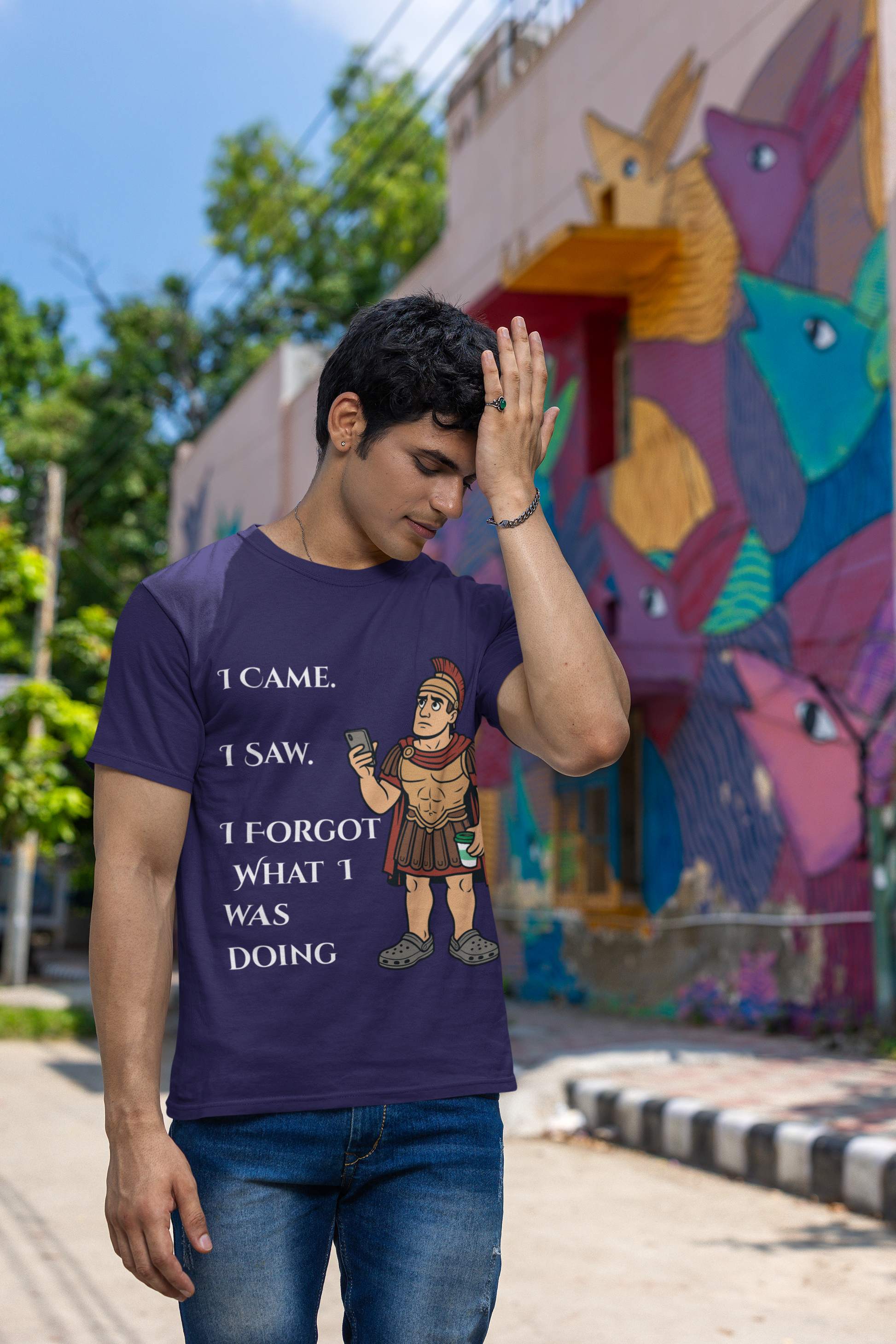 a man wearing a team navy I Came, I Saw, I Forgot T-Shirt – Funny ADHD t shirt from WhimSpin with text and graphics, standing in front of a colorful mural. he has his hand raised to his forehead as if he is just remembering something he forgot.