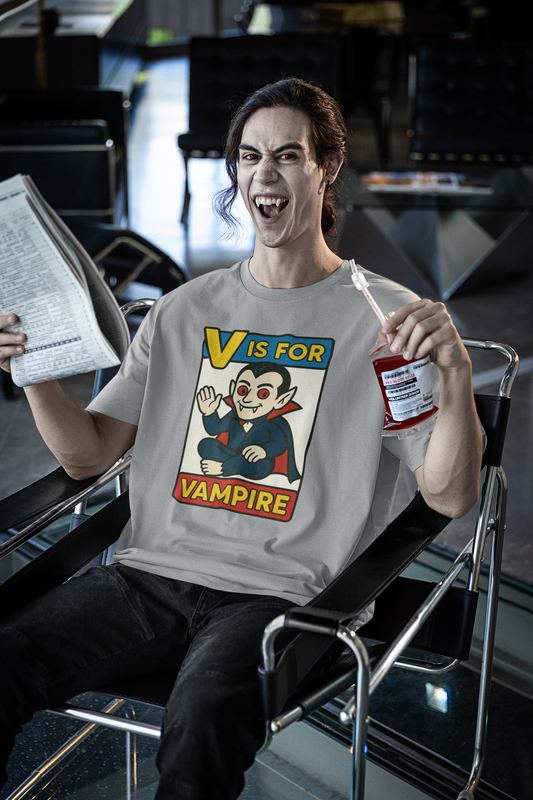 V is for Vampire T-Shirt – Funny Halloween Legend Alphabet Tee shown on a vampire-costumed man reading a newspaper and holding a blood bag in an airport, wearing the Athletic Heather color variant