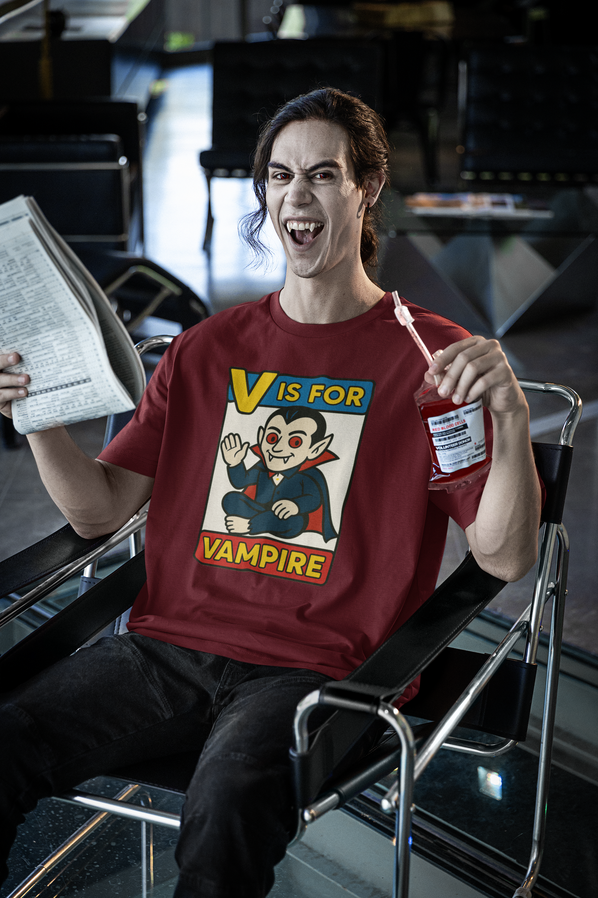 V is for Vampire T-Shirt – Funny Halloween Legend Alphabet Tee featured on a vampire man in an airport lounge, holding a blood pouch, wearing the Heather Cardinal color version