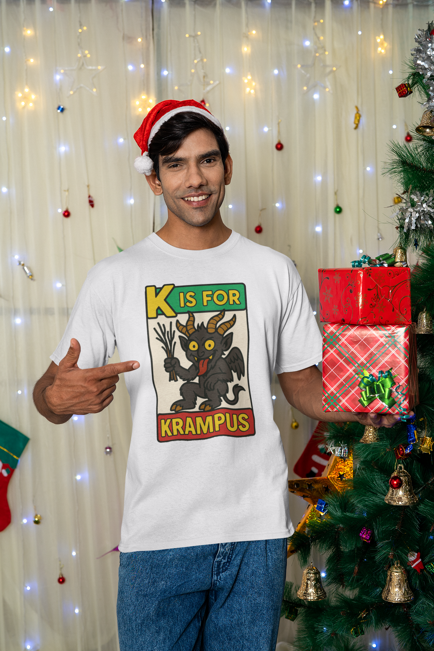 K is for Krampus Alphabet Holiday Graphic T-Shirt in White worn by man pointing at shirt in front of twinkling lights