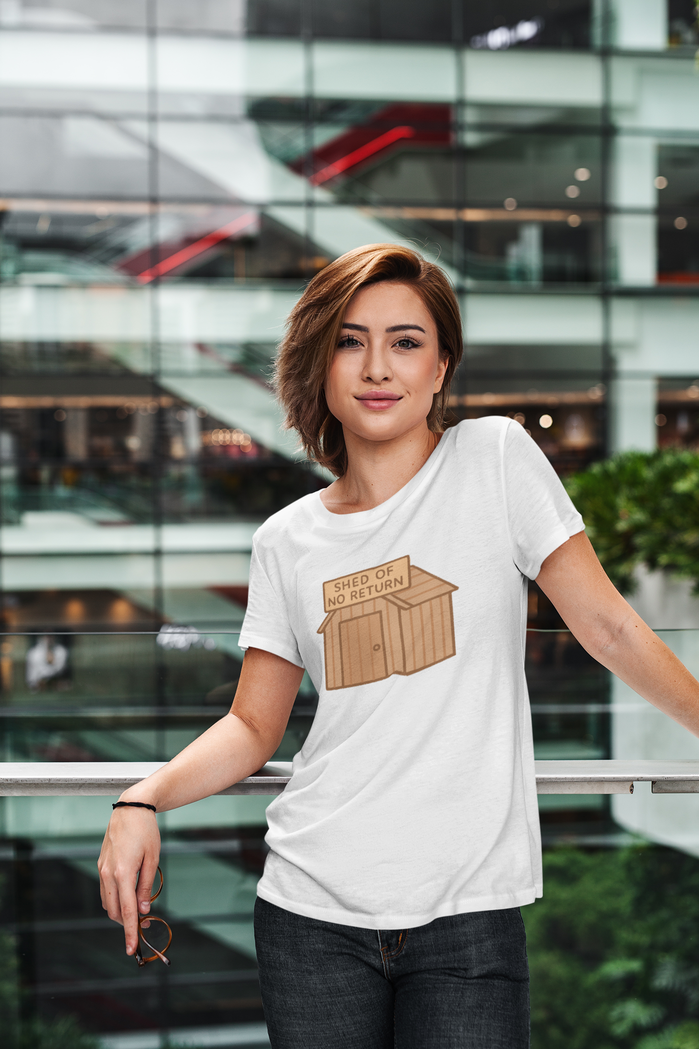 Woman wearing a white t-shirt with the "shed of no returns" graphic  stands in front of a modern glass building.
