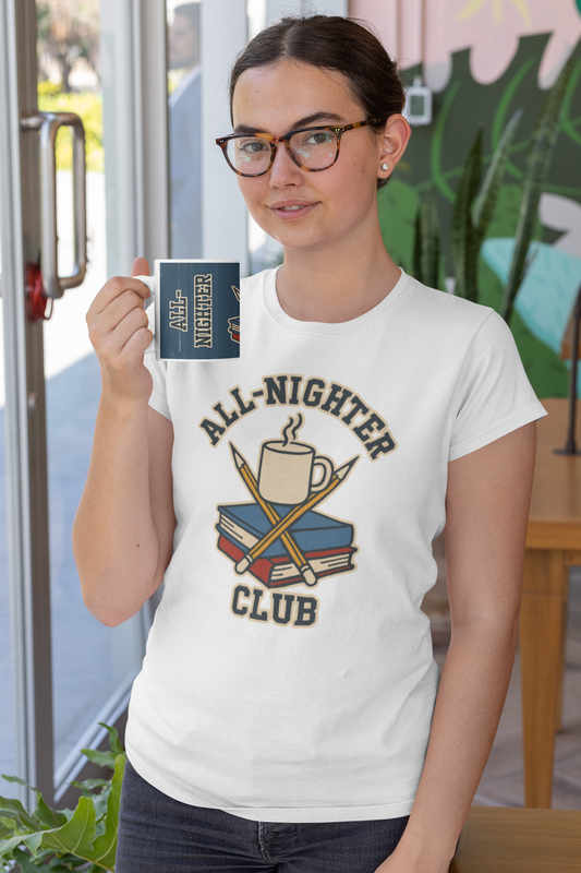 Lifestyle image of a woman in a café wearing the All-Nighter Club T-shirt in white while holding the matching mug in blue, featuring crossed pencils, books, and coffee graphic.
