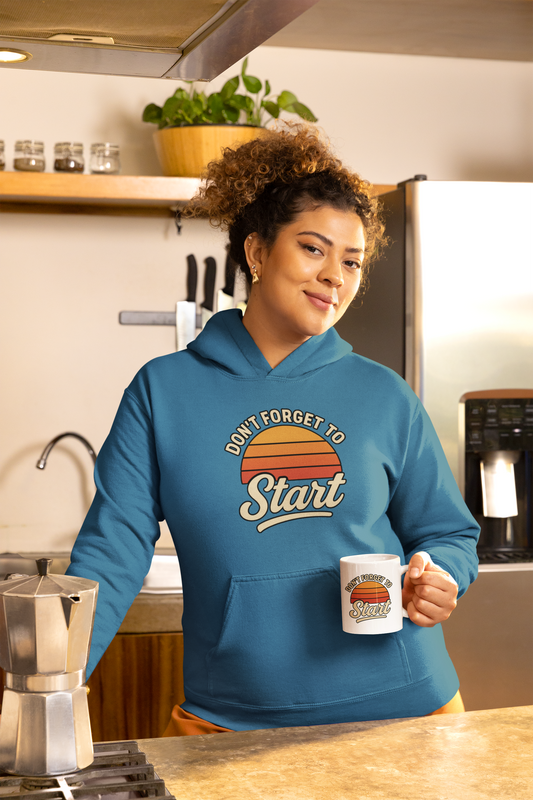 Don’t Forget to Start Hoodie – Retro Sunrise Motivational Sweatshirt worn by a woman in a kitchen holding a matching mug, highlighting the retro sunrise graphic and cozy everyday motivation.