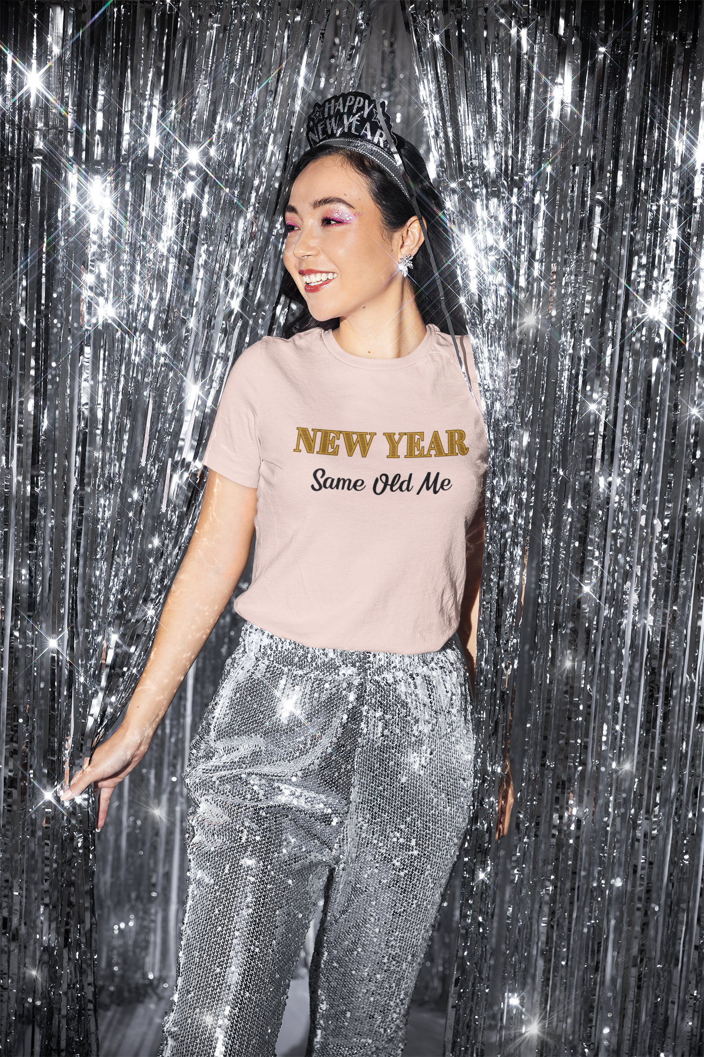 Image of a woman wearing the New Year Same Old Me T-Shirt – Funny Self-Confidence Tee with a festive headband and sequined pants, smiling against a silver tinsel backdrop.