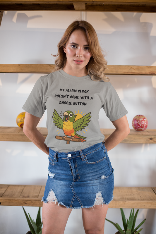 A woman wearing the Parrot Alarm Clock T-Shirt – Funny Parrot Wake-Up Call Tee in Athletic Heather, showing the shouting cartoon parrot artwork and humorous “no snooze button” message while she stands indoors against wooden shelving.