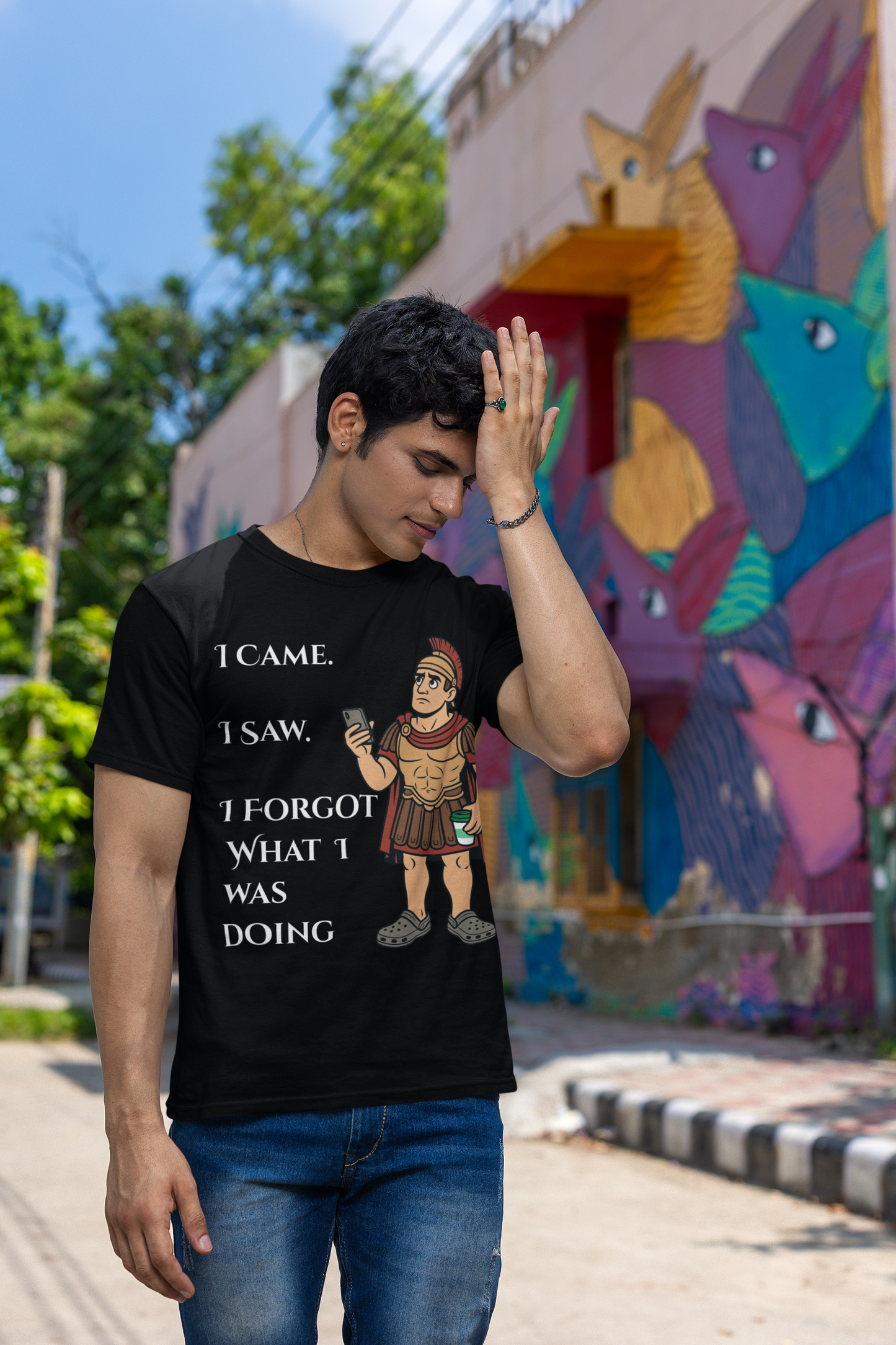 a man wearing a black I Came, I Saw, I Forgot T-Shirt – Funny ADHD t shirt from WhimSpin with text and graphics, standing in front of a colorful mural. he has his hand raised to his forehead as if he is just remembering something he forgot.